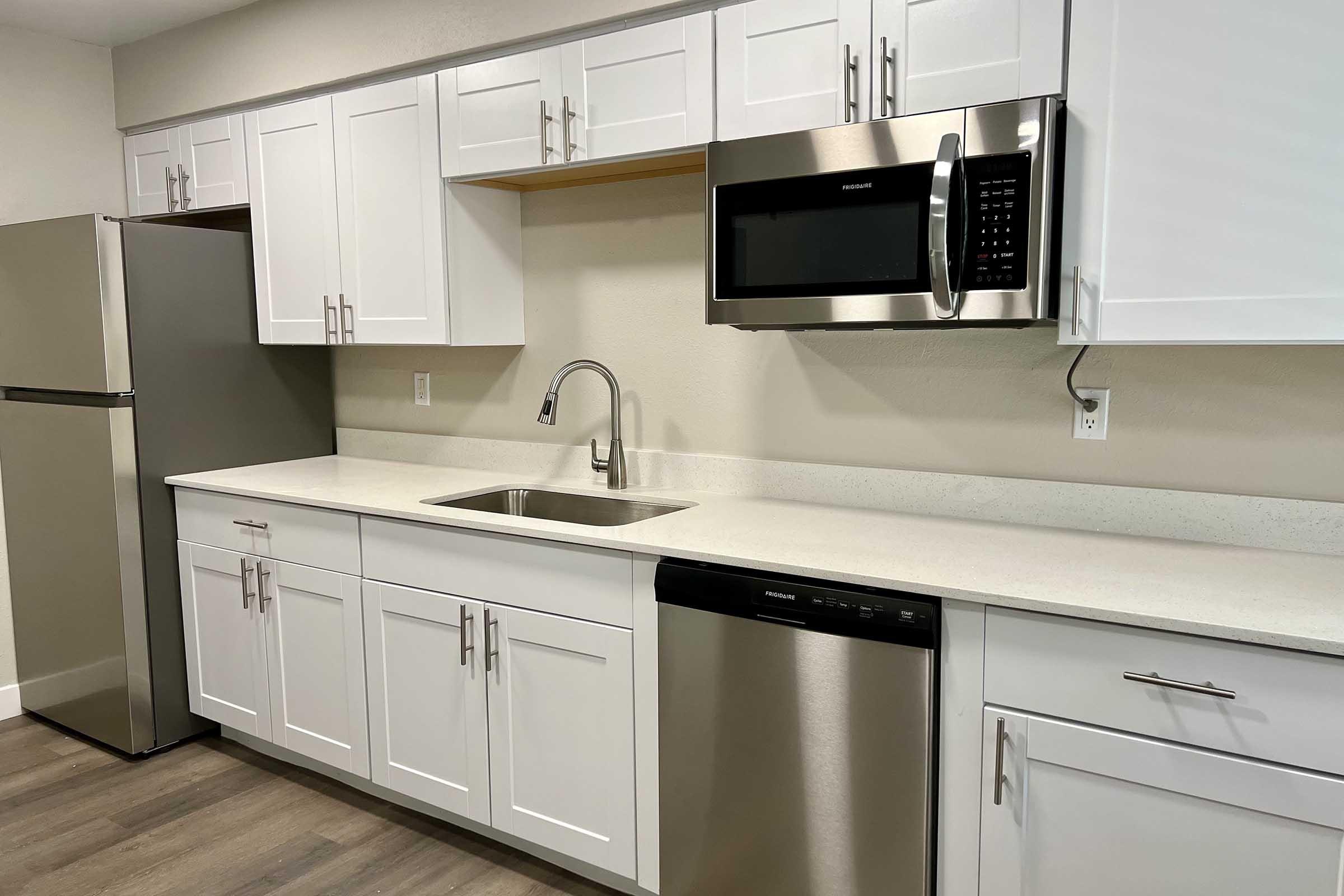 Modern kitchen featuring white cabinetry, a stainless steel refrigerator, a built-in microwave, a dishwasher, and a sink with a sleek faucet. The countertop is light-colored, and the floor has a wood-like finish, providing a clean and contemporary look.