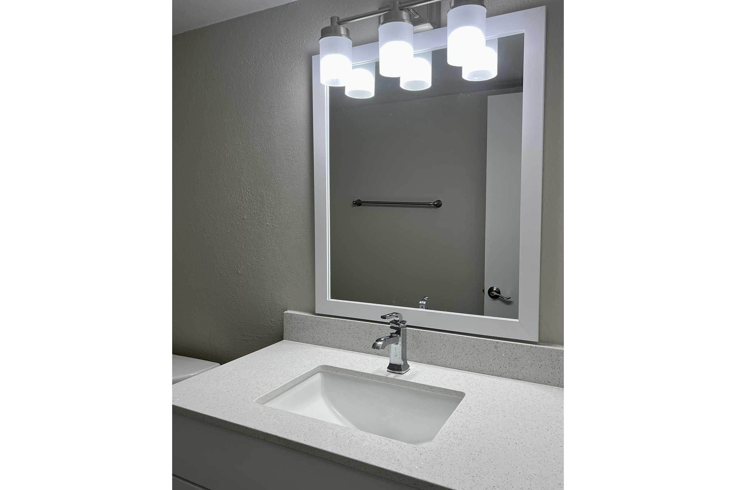 A modern bathroom sink area featuring a clean, white countertop with an integrated sink. Above the sink, there is a rectangular mirror framed in white, illuminated by three circular light fixtures. The wall is a neutral color, and a towel bar is visible in the background.
