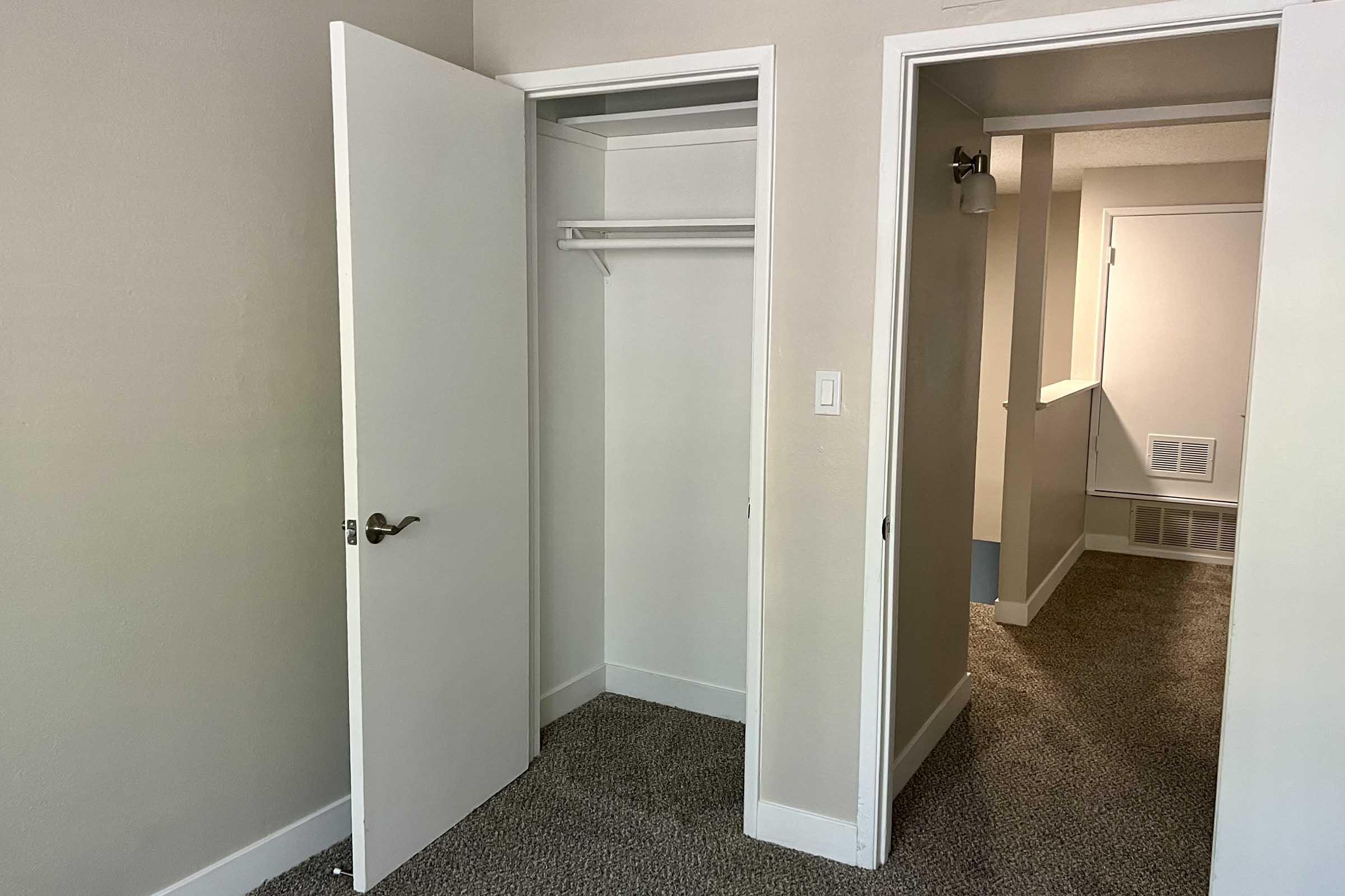 A partially furnished room featuring an open closet with a hanging rod and a beige wall. A doorway leads to another area, suggesting a small hallway with light-colored walls and a textured carpet floor. Natural light illuminates the space, creating a clean and simple aesthetic.