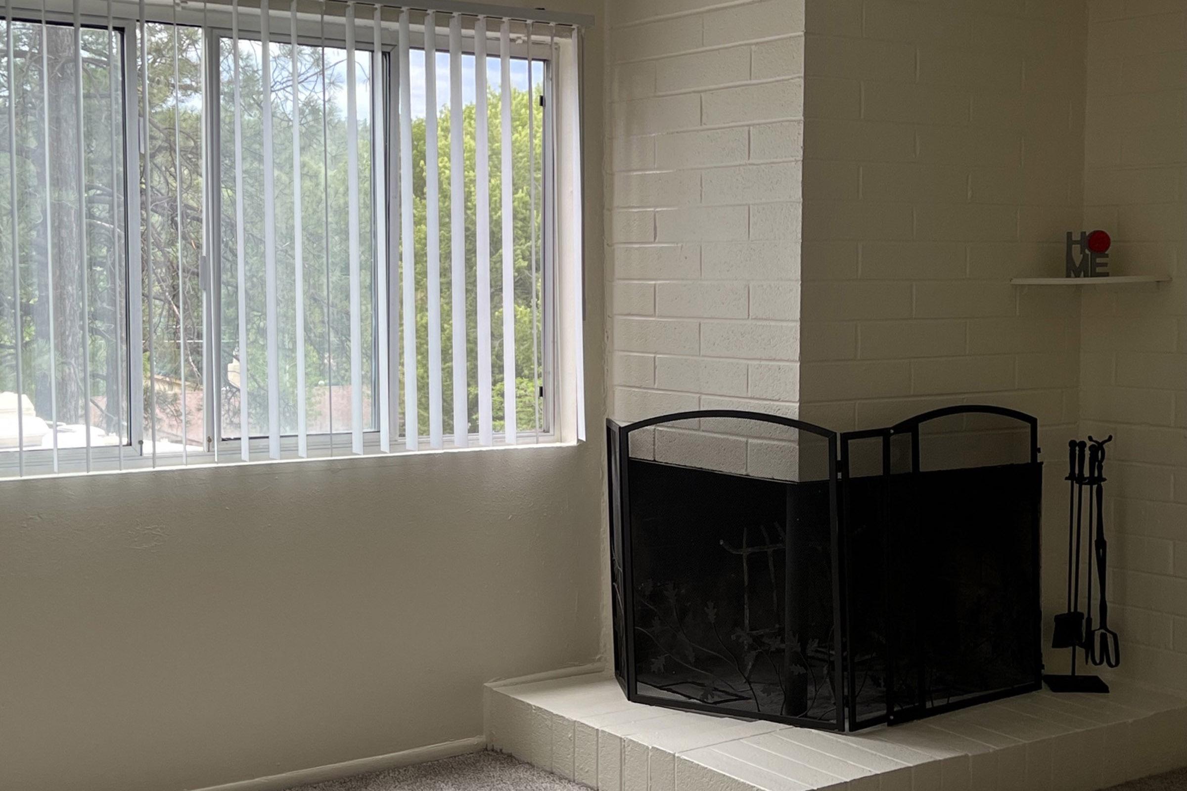 A cozy living room featuring a white brick fireplace with a black screen and tools. Large vertical blinds cover a window, allowing soft light to illuminate the space. Outside, greenery is visible, enhancing the inviting atmosphere of the room. A small shelf with the word "HOME" adds a personal touch.