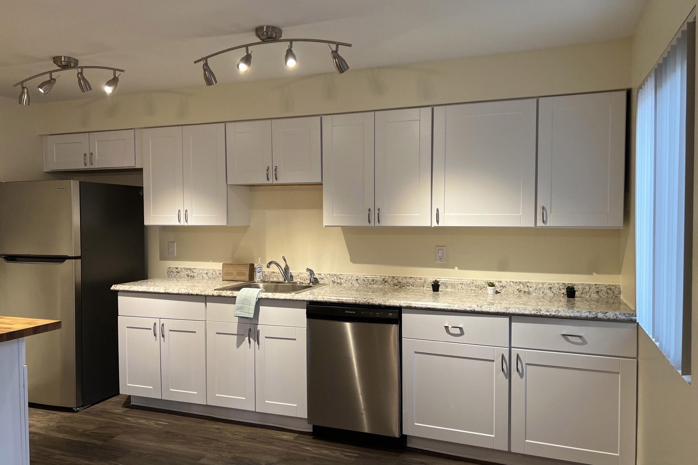 A modern kitchen with white cabinetry, stainless steel appliances including a refrigerator and dishwasher, and a granite countertop. The space features track lighting and neutral-colored walls, with a window providing natural light.