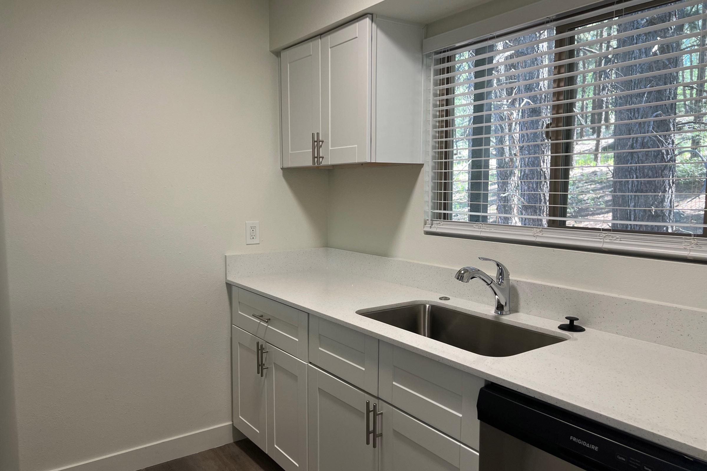 Modern kitchen with white cabinetry, a stainless steel sink, and a countertop made of light-colored material. A window with blinds provides natural light, revealing greenery outside. The flooring is a light wood finish, enhancing the contemporary look of the space.