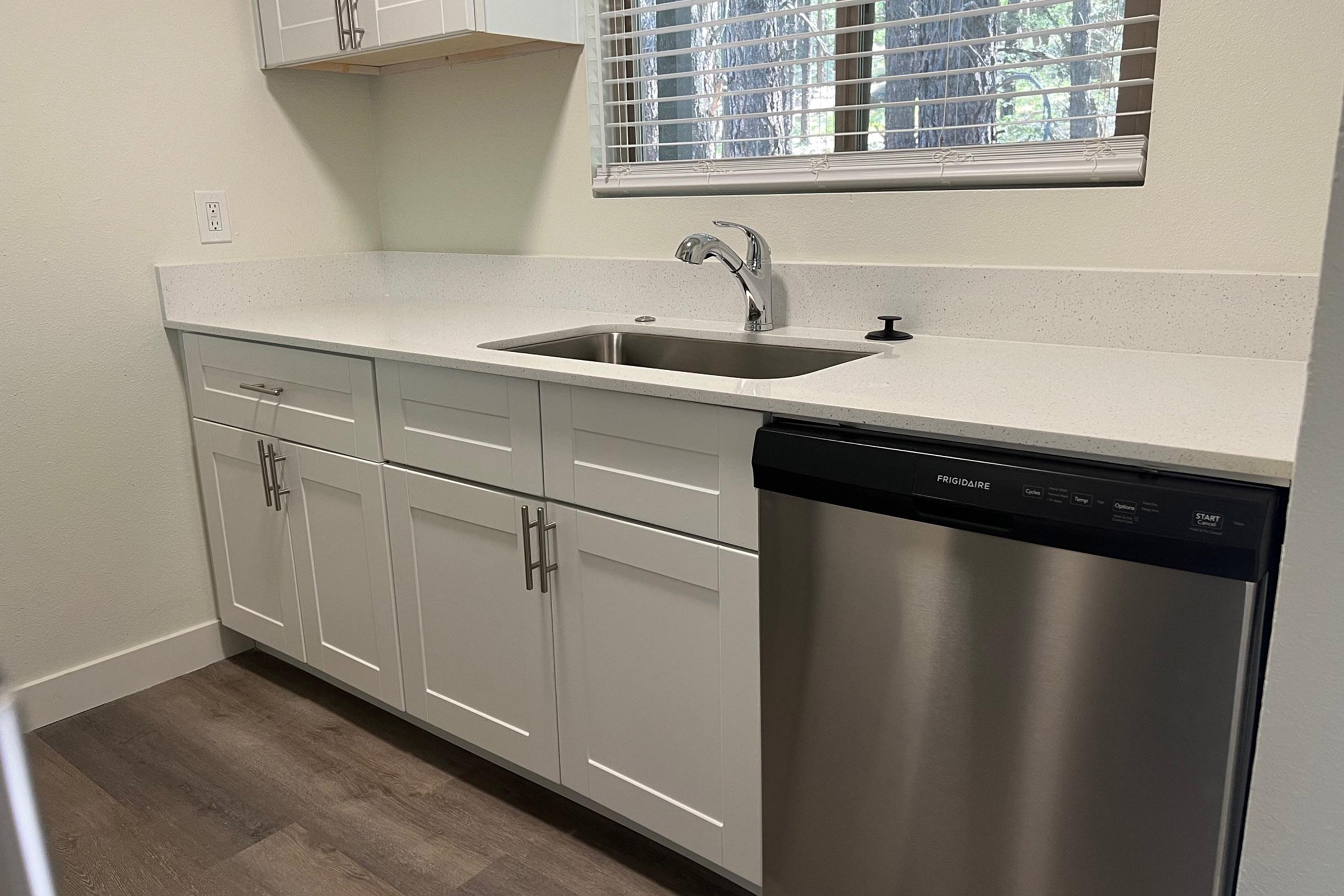 Modern kitchen featuring a sleek countertop with a stainless steel sink and faucet. Below, white cabinets are paired with a silver dishwasher, set against a backdrop of neutral walls and natural light streaming in through a window. The floor is finished with wood-like flooring.