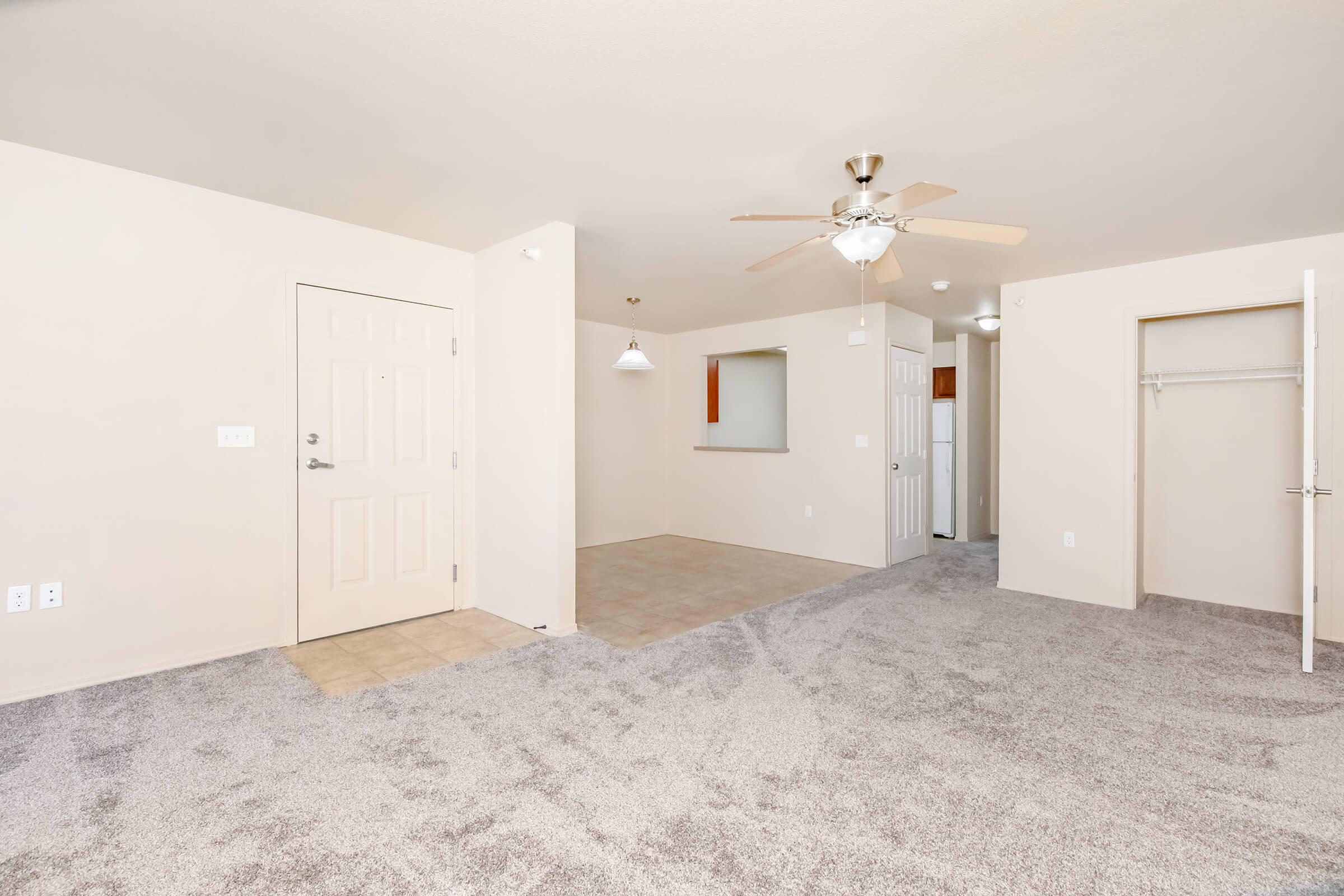 Spacious living area of an apartment featuring light-colored walls, plush carpet, a ceiling fan, and a doorway leading to a closet. The layout includes an open space with a small kitchen counter visible in the background, and a door to the exterior. Natural light brightens the room.
