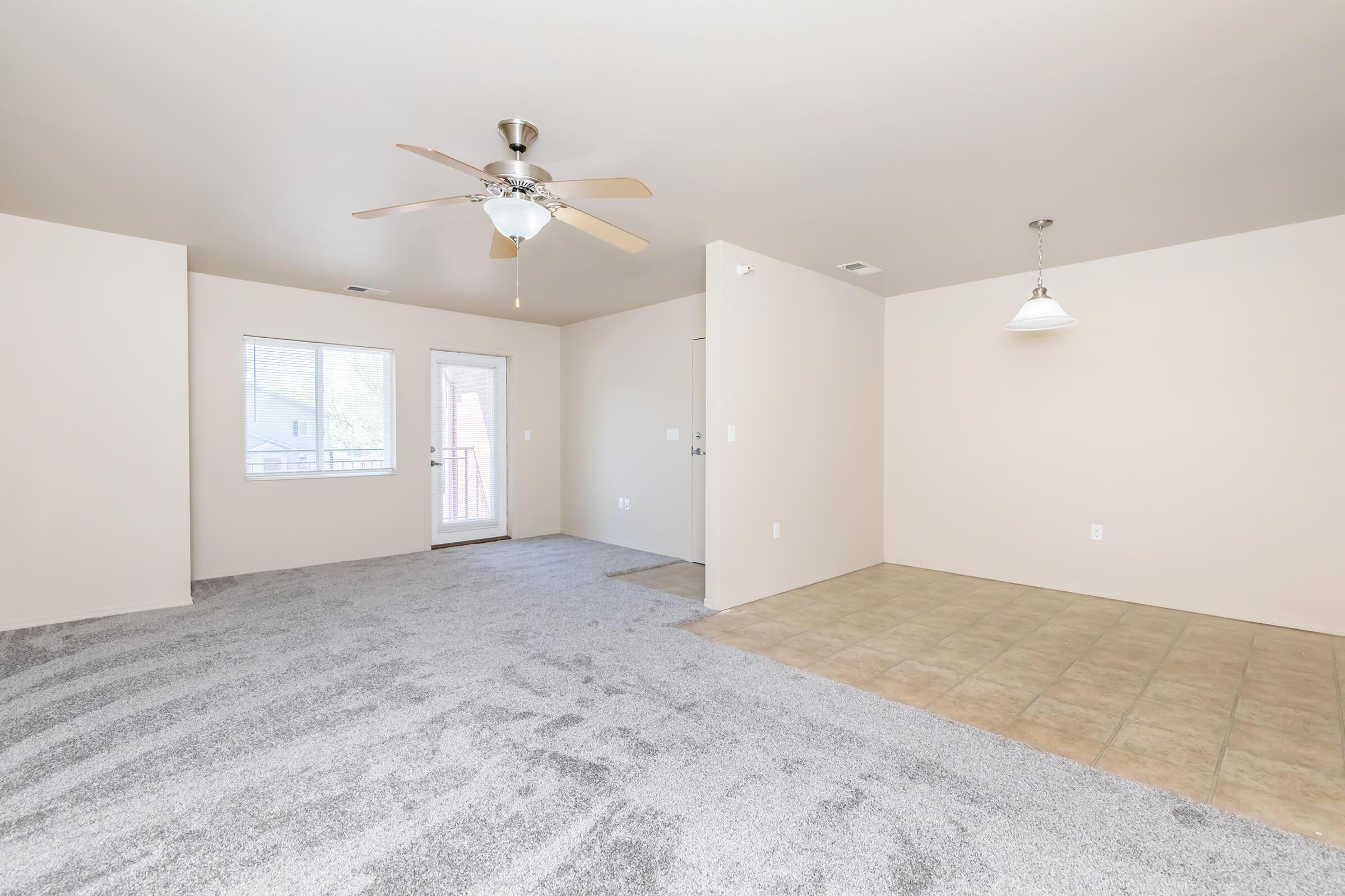 A spacious, well-lit living area featuring a ceiling fan, carpeted flooring, and a light-colored wall. To the left, a door leads outside, and a window allows natural light to enter. The adjacent area has tiled flooring, suitable for a dining space or additional furniture.