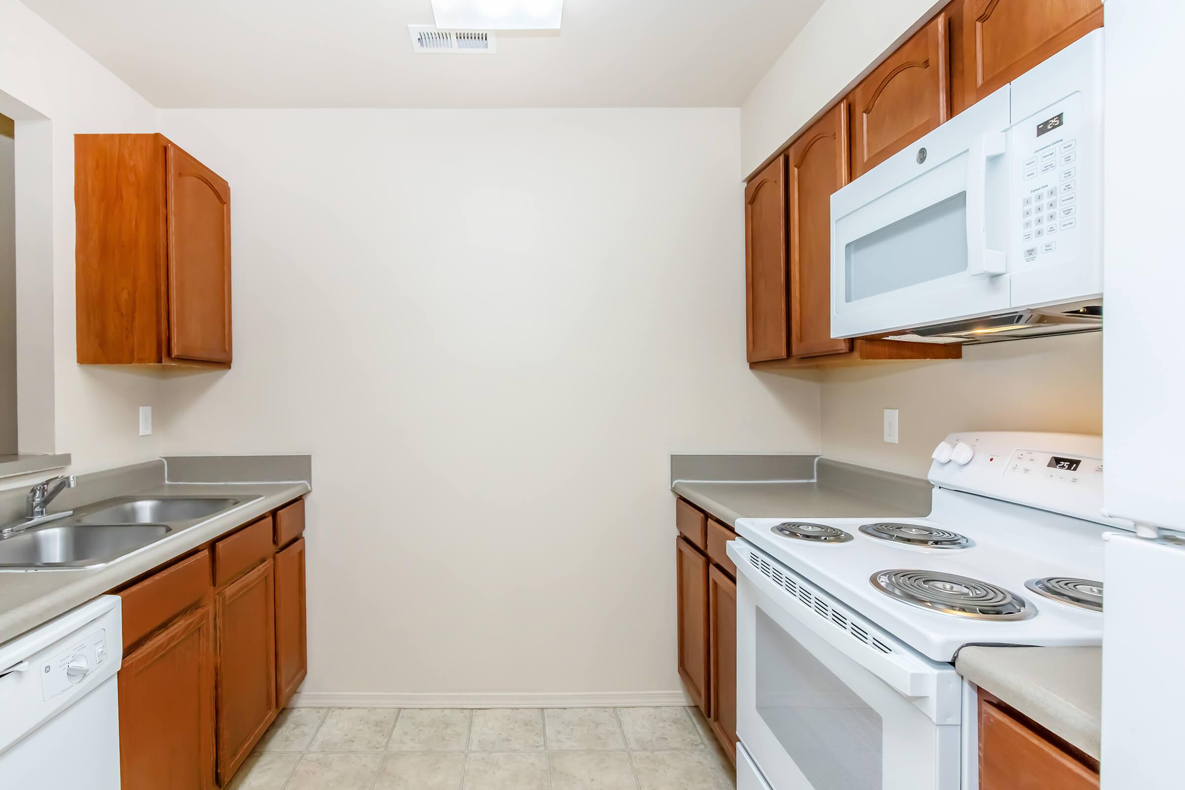 A view of a kitchen with wooden cabinets, a white microwave above the stove, and a sink on the left. The kitchen features gray countertops and a light-colored tile floor, creating a clean and simple design. The walls are painted a neutral shade, enhancing the spacious feel of the area.