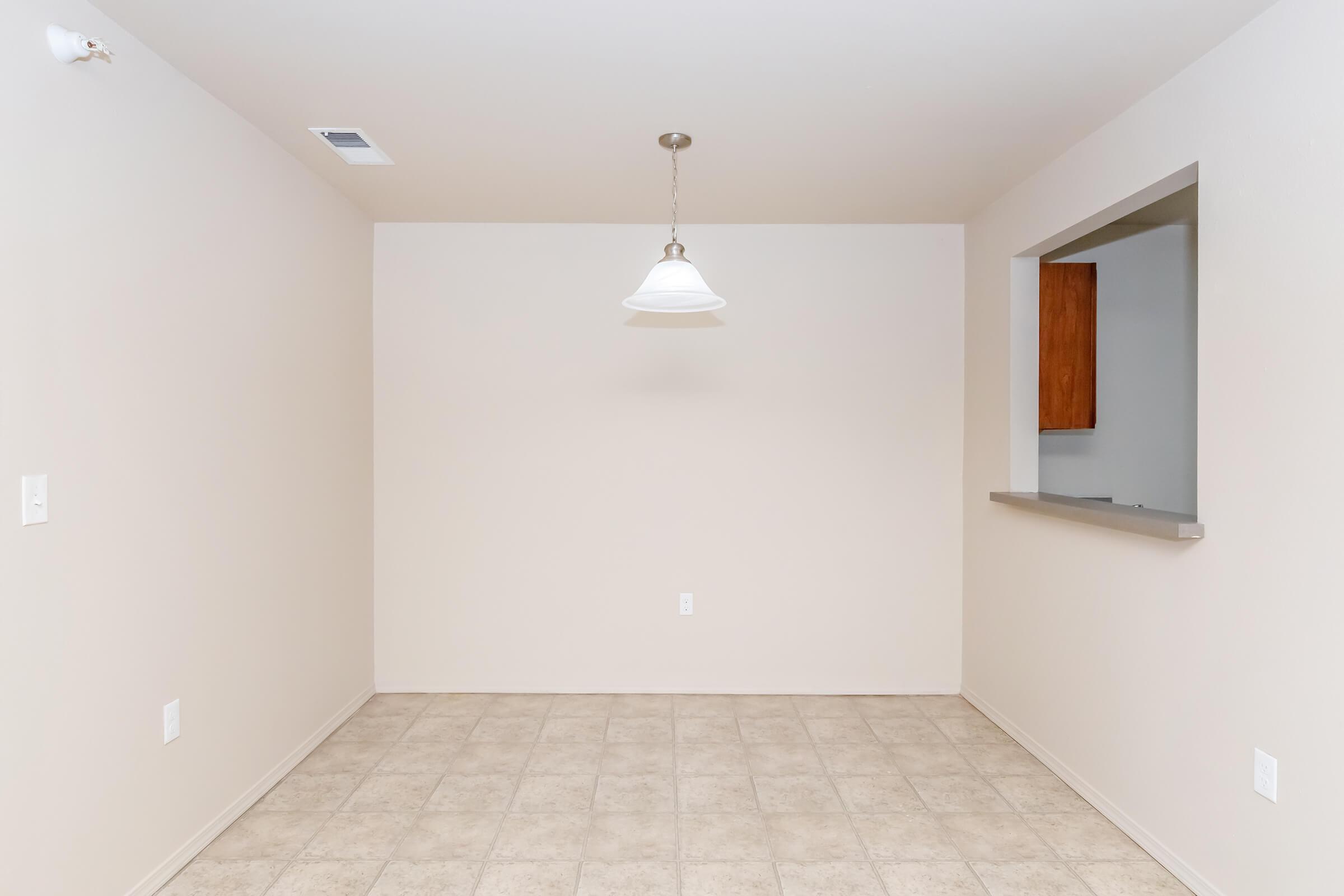 A vacant dining area with beige walls and tiled floor. A single pendant light hangs from the ceiling, and there's a pass-through window to a kitchen area. The space is empty and bright, giving a sense of openness.