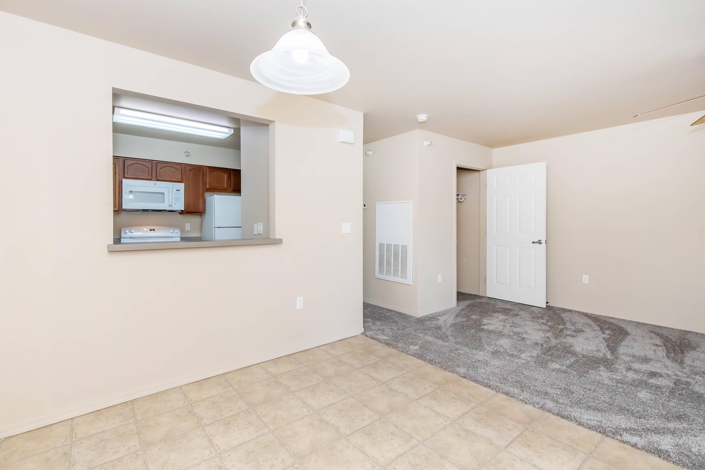 An interior view of an apartment featuring a beige wall color, a window into the kitchen showcasing wooden cabinets, a white microwave, and a refrigerator. The living area has light-colored tiled flooring and a carpeted space with a door leading to another room. A light fixture hangs from the ceiling.