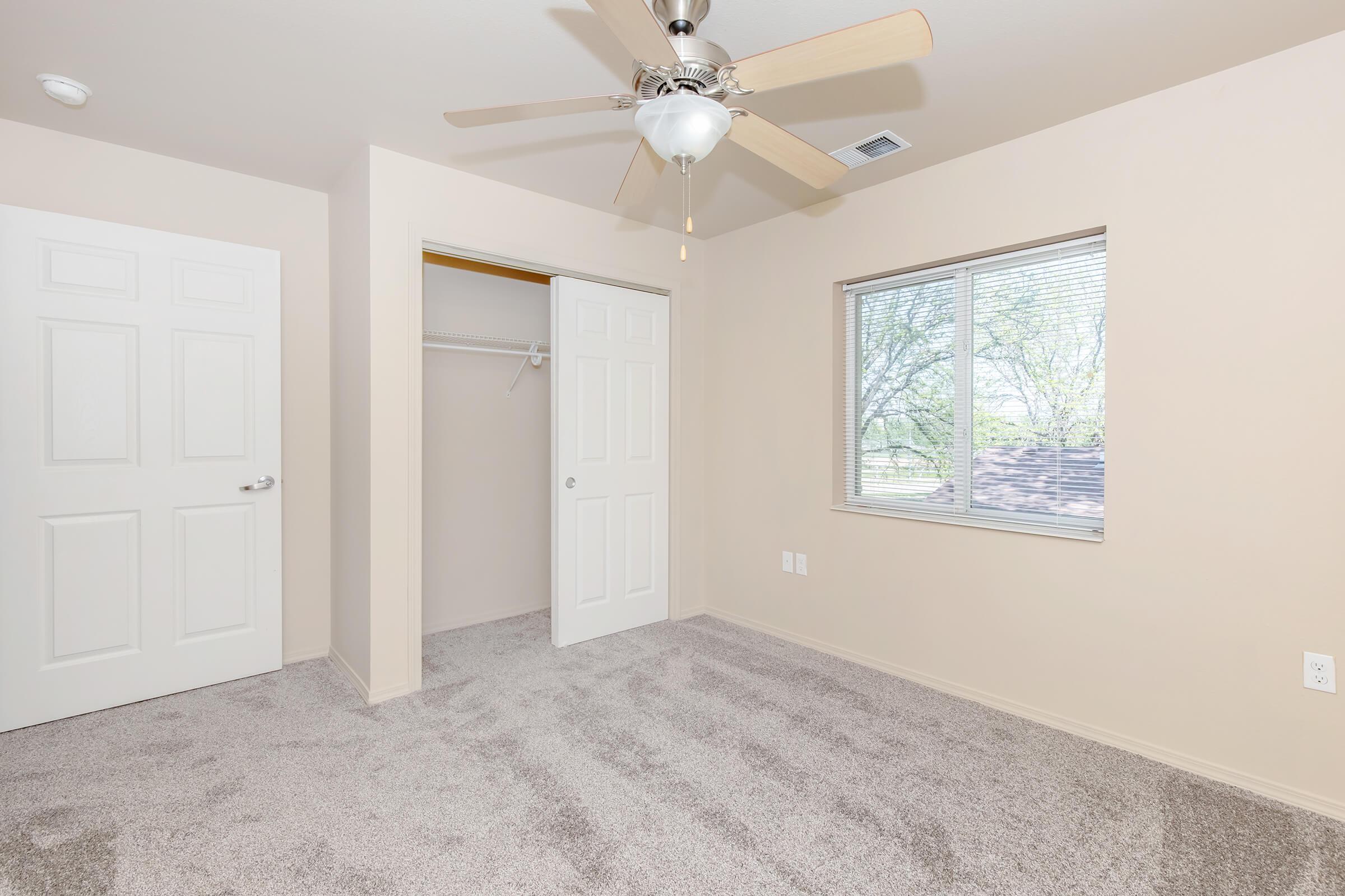 A clean, empty bedroom with light beige walls, a ceiling fan, and a window letting in natural light. There are two white doors, one leading to a closet, and soft carpet covering the floor. The room appears spacious and inviting, ideal for personal decoration.