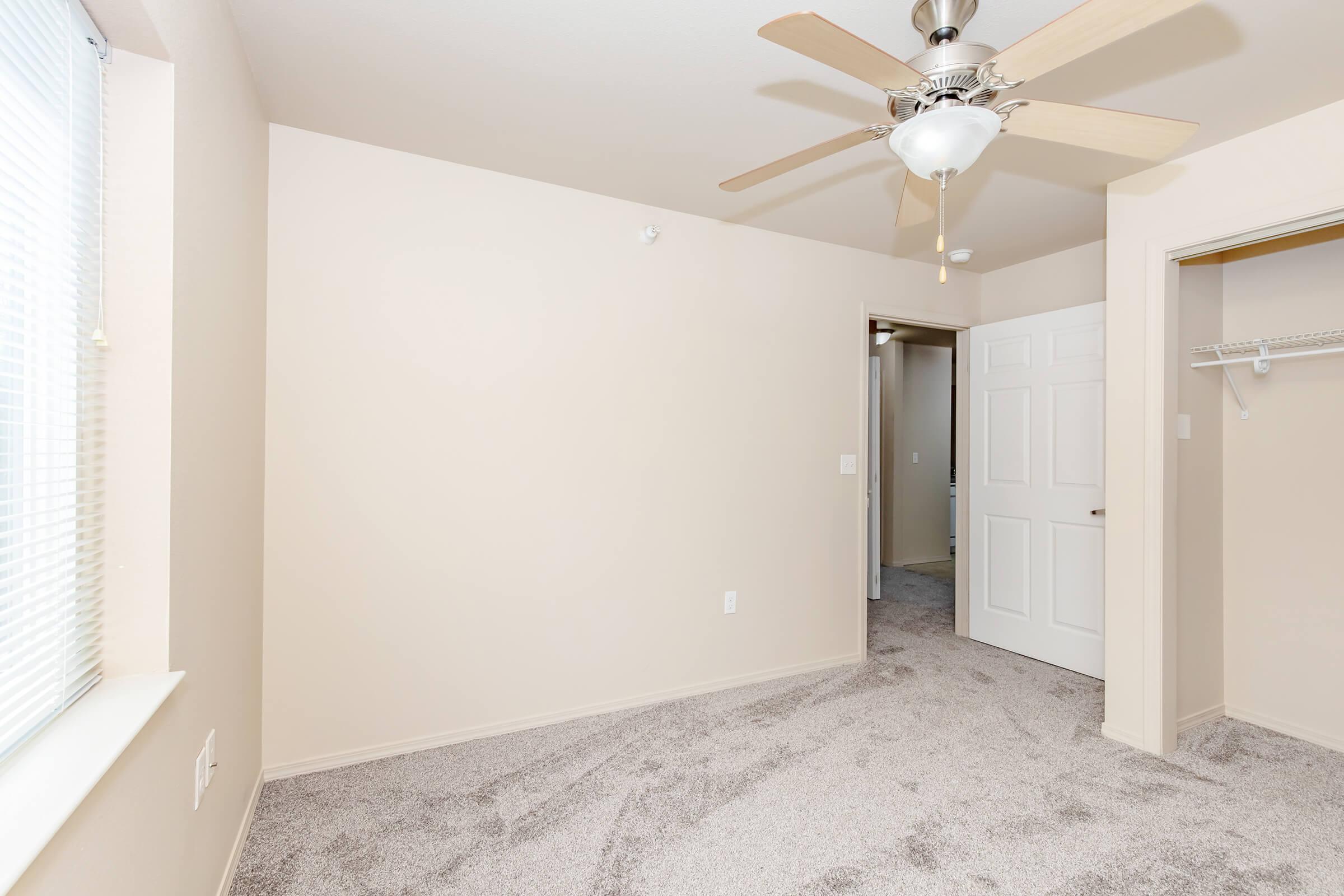 A light-colored room with beige walls and soft gray carpeting. A ceiling fan with light hangs from the ceiling. One wall features a window with sheer blinds, and there is a doorway leading to another room. A closet space is visible on the right. The room appears bright and spacious.