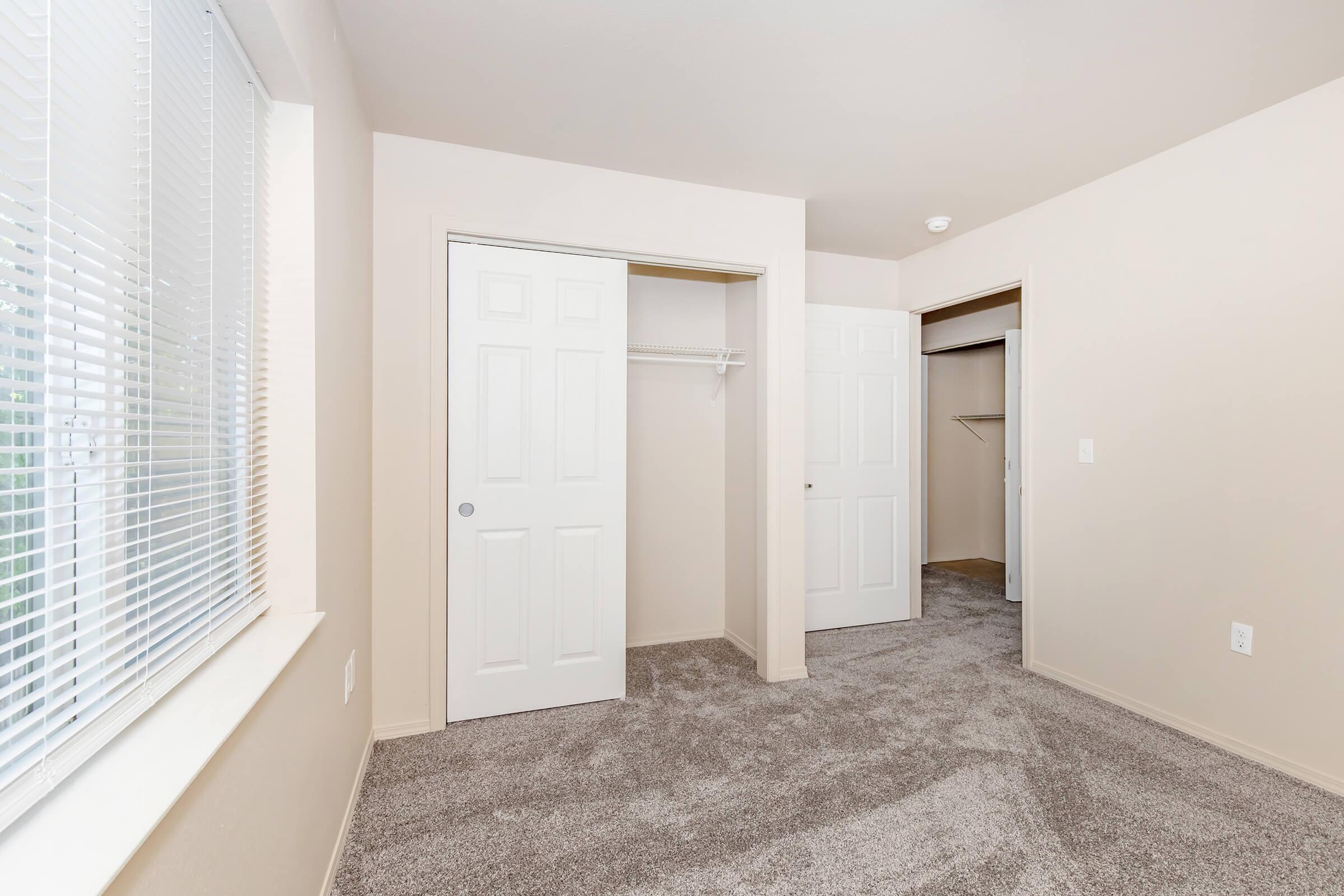 A small, empty room with beige walls and carpeted floor. There are two doors leading to closets, one with a hanging rod visible. The room features a large window with blinds, allowing natural light to enter, creating a bright and airy atmosphere.