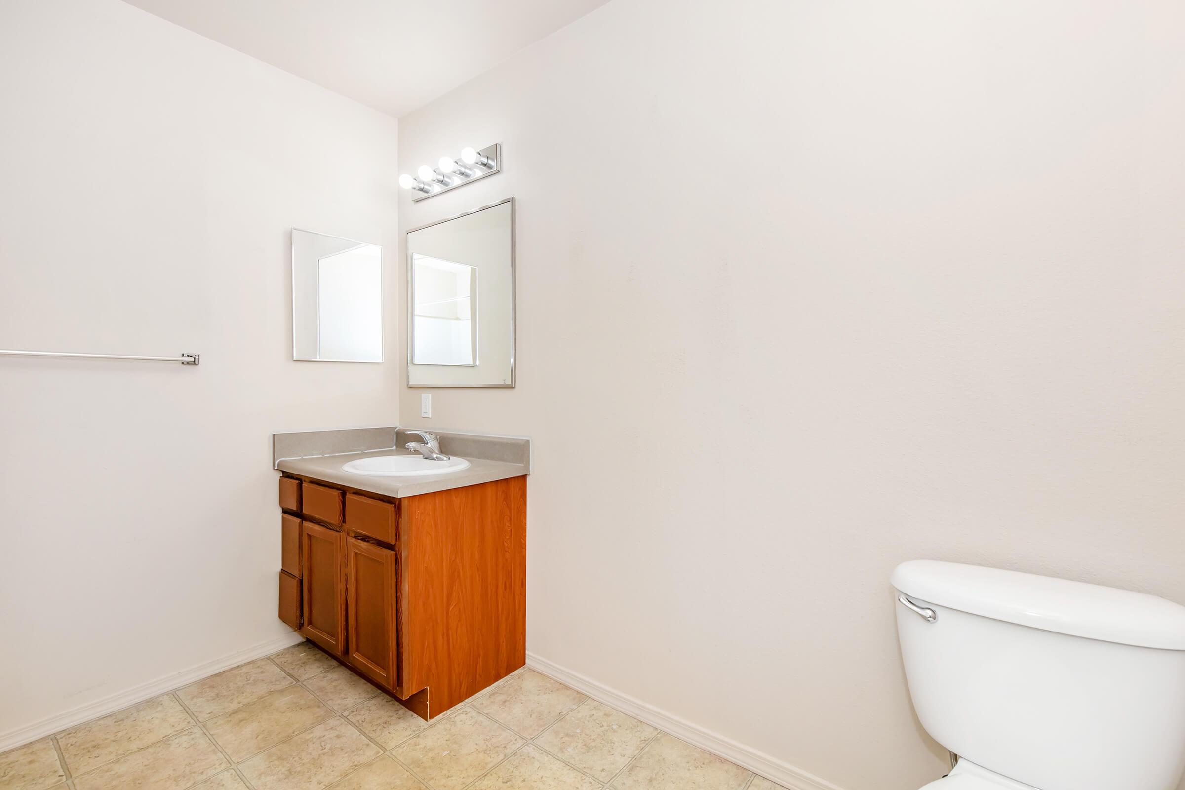 A clean, minimalist bathroom featuring a light-colored wall, a single sink vanity with a mirror above, wooden cabinetry, and a white toilet. The floor is tiled in a neutral tone, and there is ample empty wall space, creating an open and airy feel.