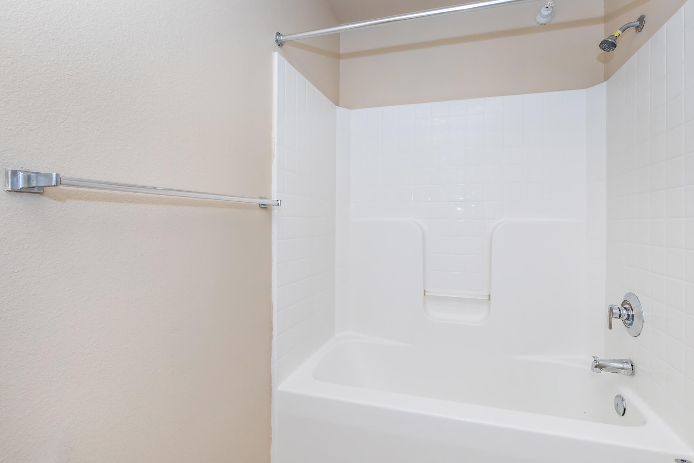 A clean, modern bathtub with a white tiled surround and showerhead, featuring a chrome grab bar on the left and a chrome faucet. The bathroom walls are a light color, creating a bright and airy atmosphere.