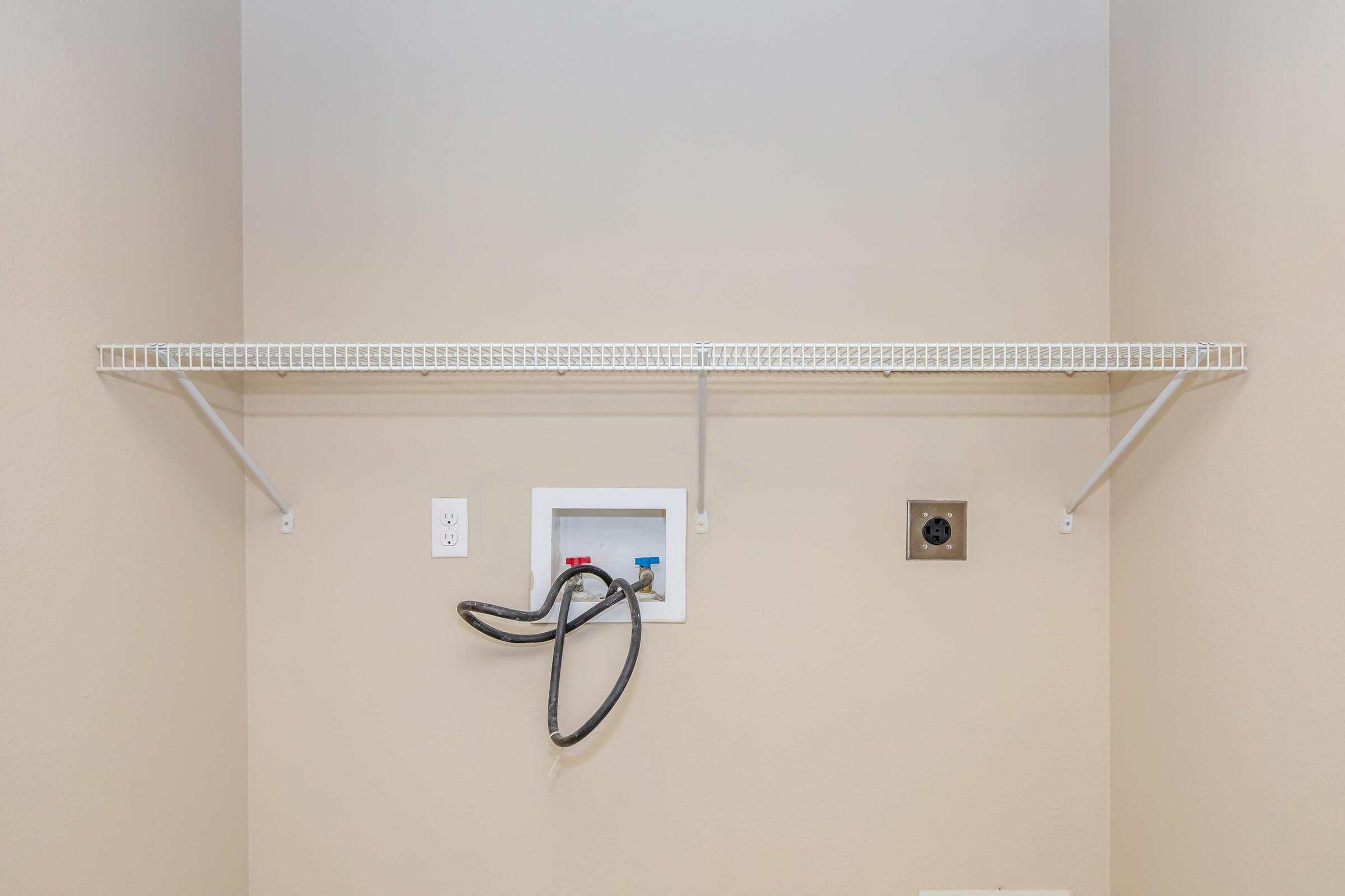 A clean, empty laundry room featuring a beige wall, a wire shelf mounted above, and a wall-mounted connection panel for laundry appliances with hoses attached. Power outlets are visible, indicating a designated area for laundry activities.
