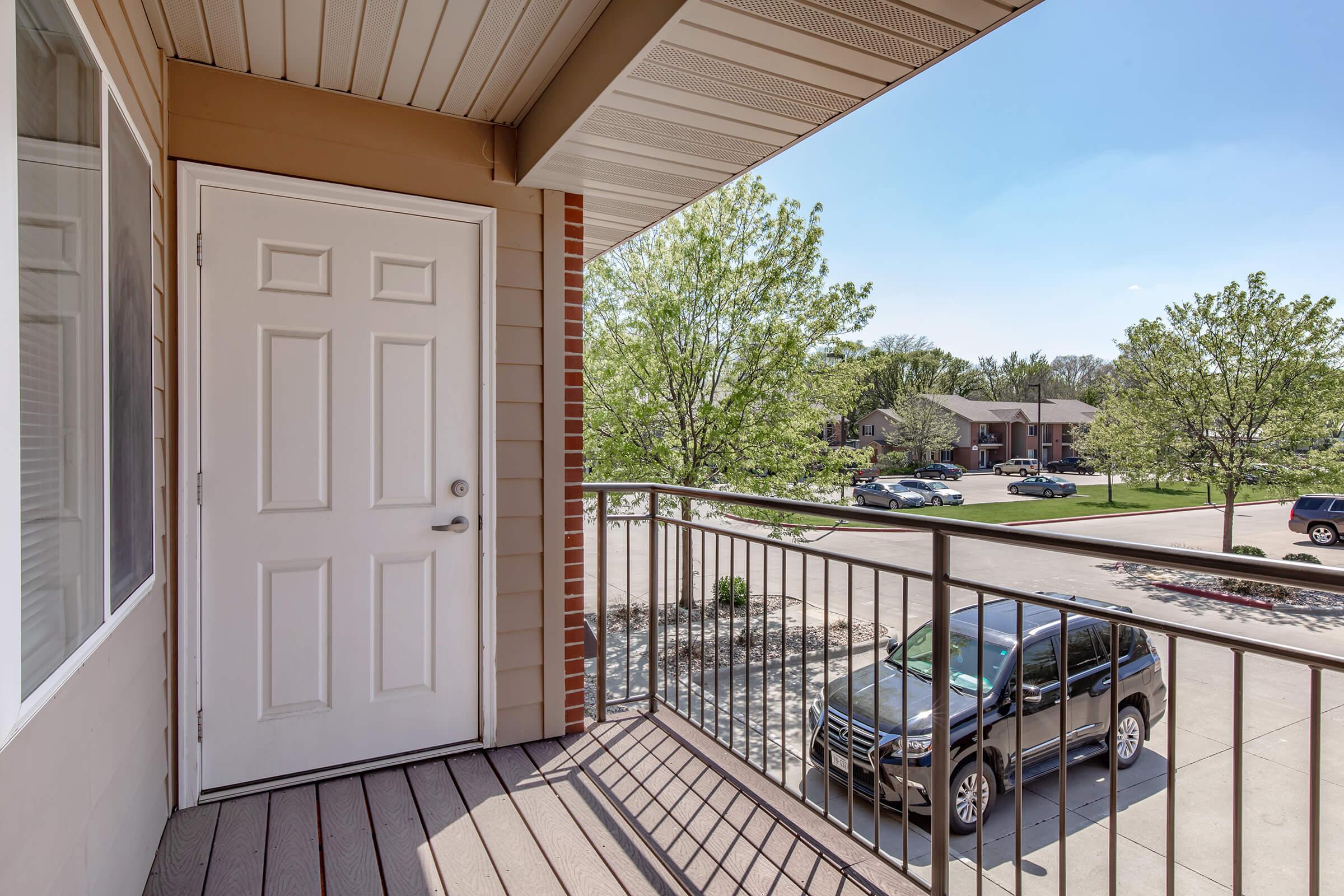 A view from a balcony featuring a closed white door, wooden flooring, and a railing. In the background, there are green trees and a parking area with several cars. The sky is clear and blue, indicating sunny weather.