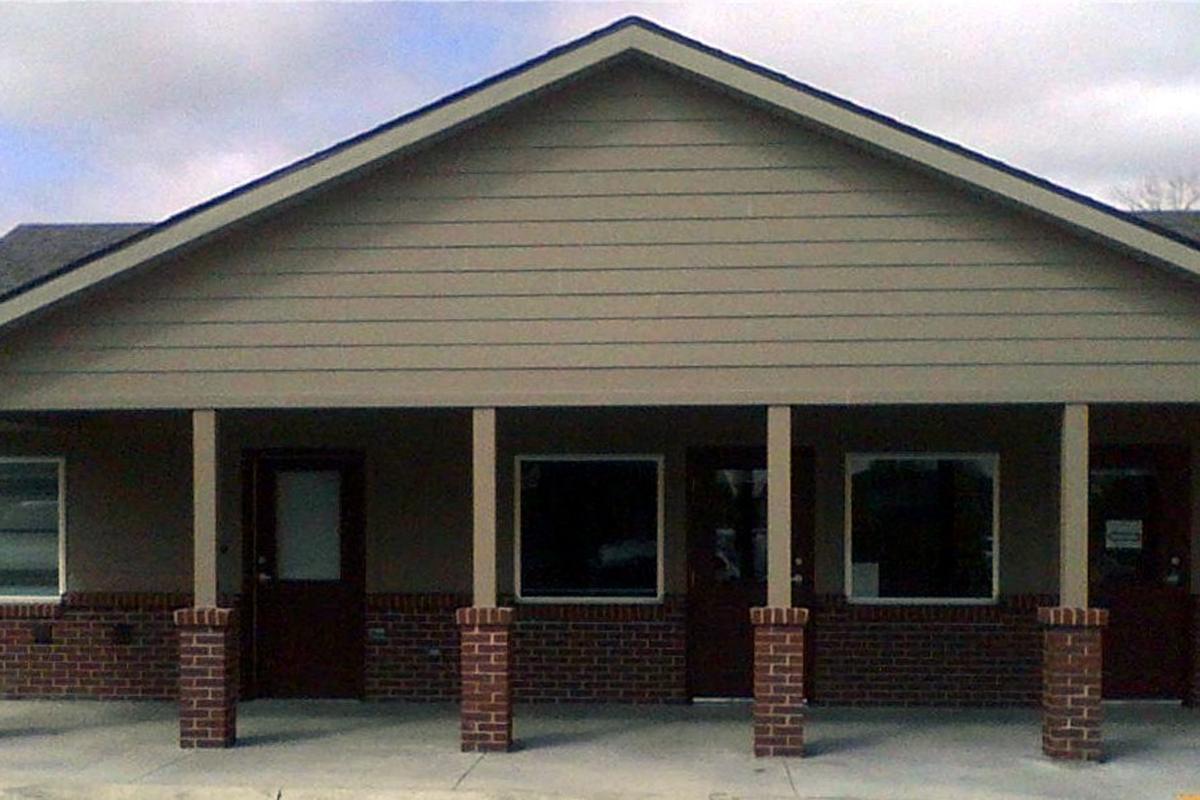 A single-story building with a sloped roof and light brown siding. The facade features four entrance doors, two on each side, with brick pillars supporting the porch area. The windows adjacent to the doors provide natural light. The sky is cloudy, creating a subdued atmosphere.