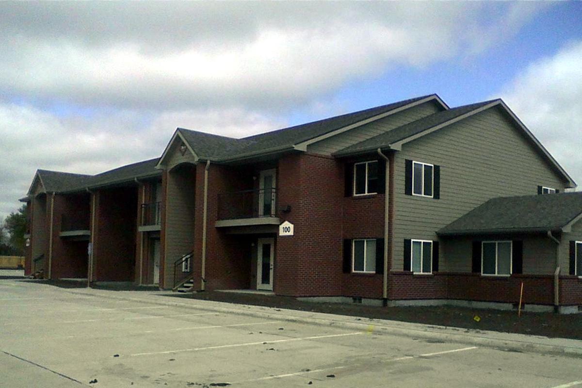 A two-story apartment building with a gray and red brick exterior. The building features multiple windows and a front entrance with stairs. The surrounding area is partially paved with a parking lot, and the sky is overcast with scattered clouds.