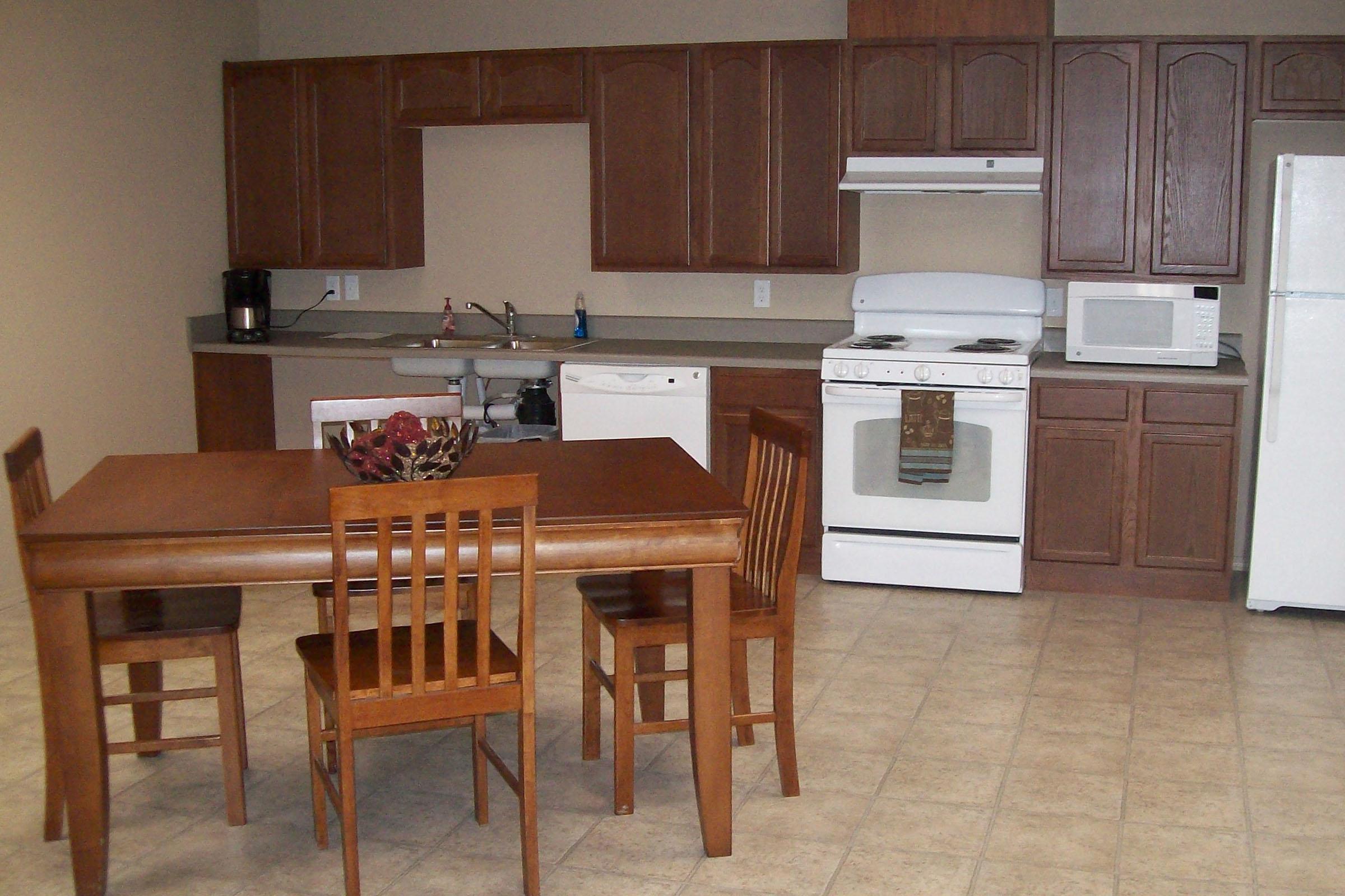 A modern kitchen featuring wooden cabinets, a countertop, and essential appliances, including a stove, refrigerator, microwave, and dishwasher. In the foreground, there is a wooden dining table with four matching chairs, and a decorative bowl at the center of the table. The floor is tiled.