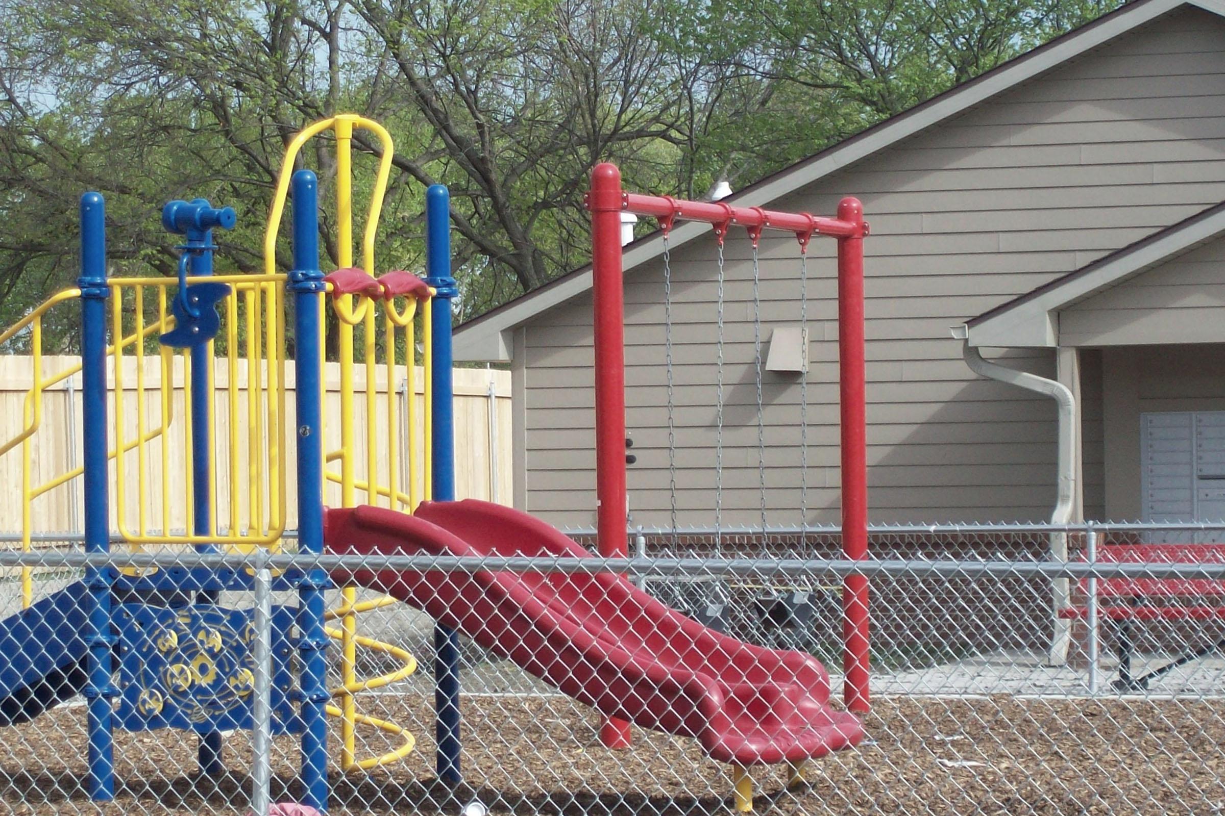 Colorful children's playground equipment featuring a yellow climbing structure and a red slide, alongside a row of swings. The playground is enclosed by a fence and is situated next to a beige house. The ground is covered with wood chips, and the scene is surrounded by trees.