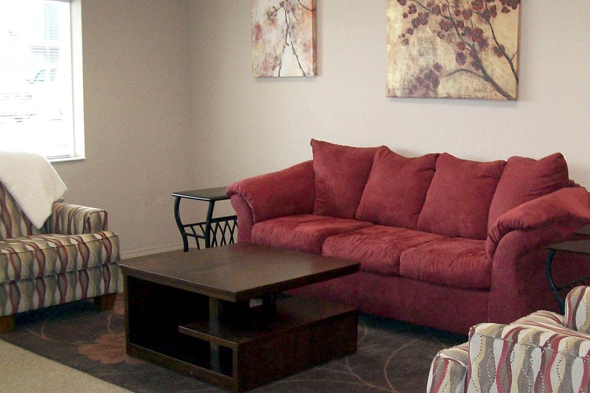 A cozy living room with a red sofa and two striped armchairs. There's a dark coffee table in the center, surrounded by a patterned rug. Two floral wall art pieces hang on the beige walls, and natural light comes in from a window, creating a warm, inviting atmosphere.