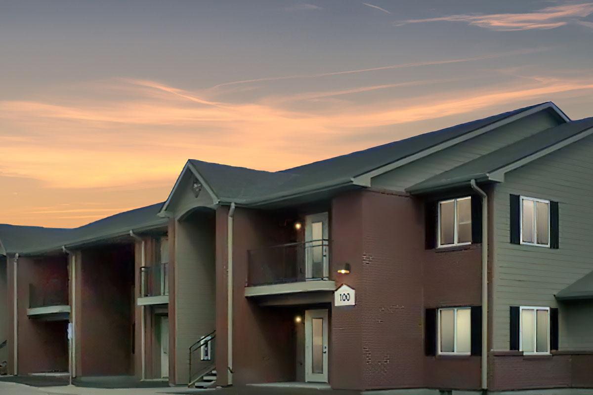 A peaceful residential building at dusk, featuring a mix of brick and siding exteriors. The entrance is highlighted by warm lighting, and the sky above is painted in soft hues of orange and purple, suggesting the approach of evening. The structure has a modern design with a welcoming atmosphere.