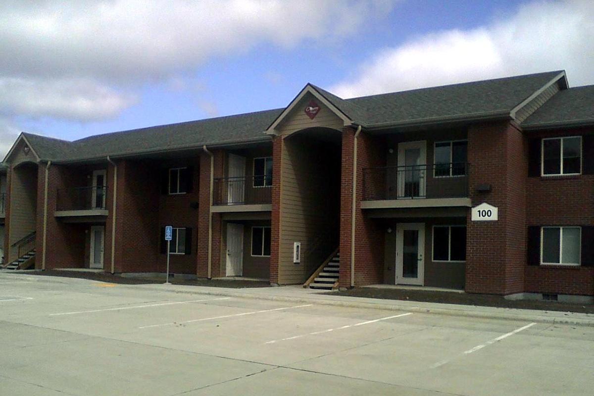 View of a two-story brick apartment building featuring multiple entrances with balconies. The building has a peaked roof and a sign with a heart symbol above the central entrance. Parking spaces are visible in front, and the sky is partly cloudy, suggesting a clear day.