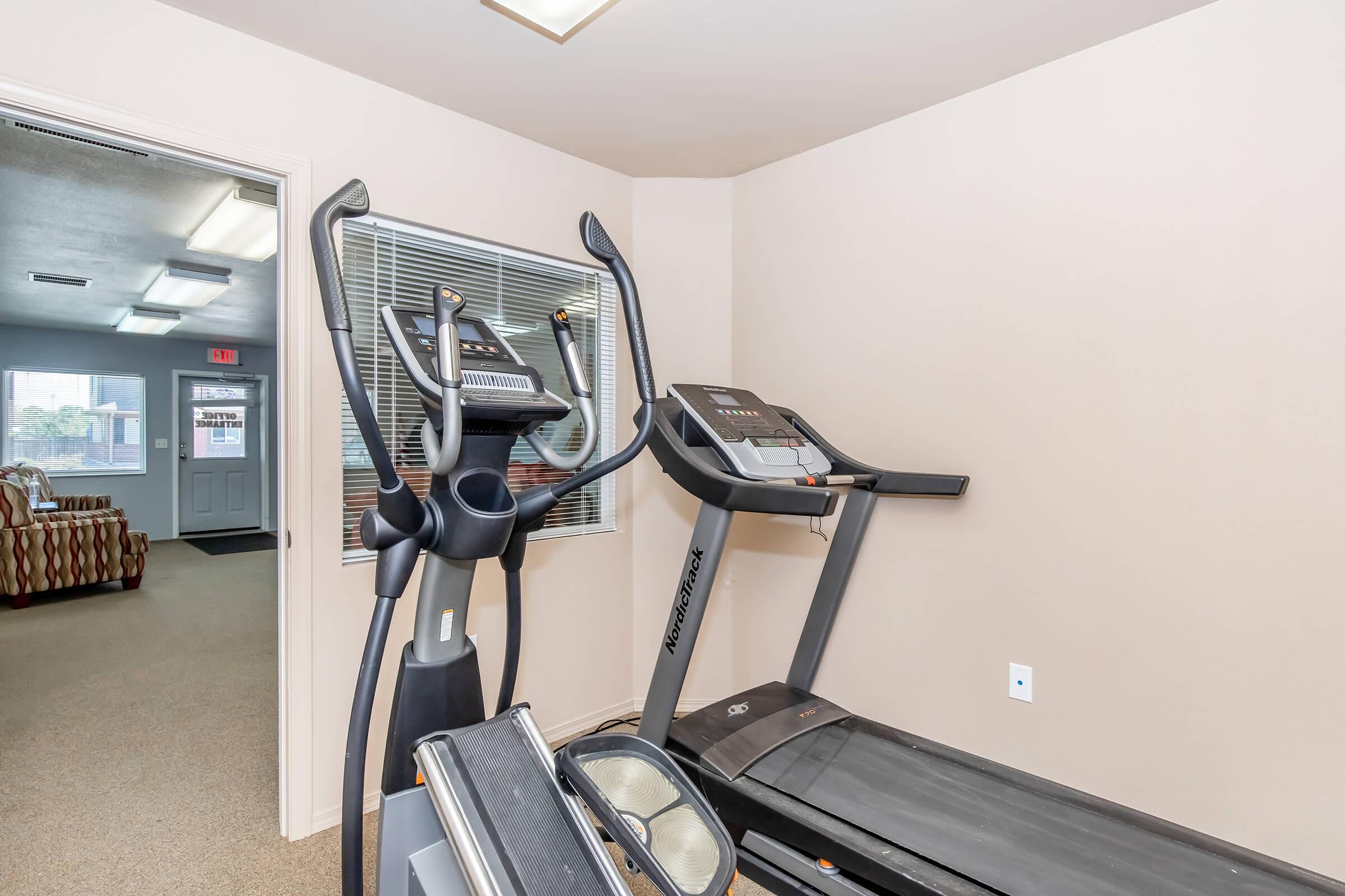 A fitness area featuring a treadmill and an elliptical machine, with a light-colored wall and a window that brings in natural light. In the background, there's a glimpse of a couch and an entrance door, suggesting a well-maintained exercise space.