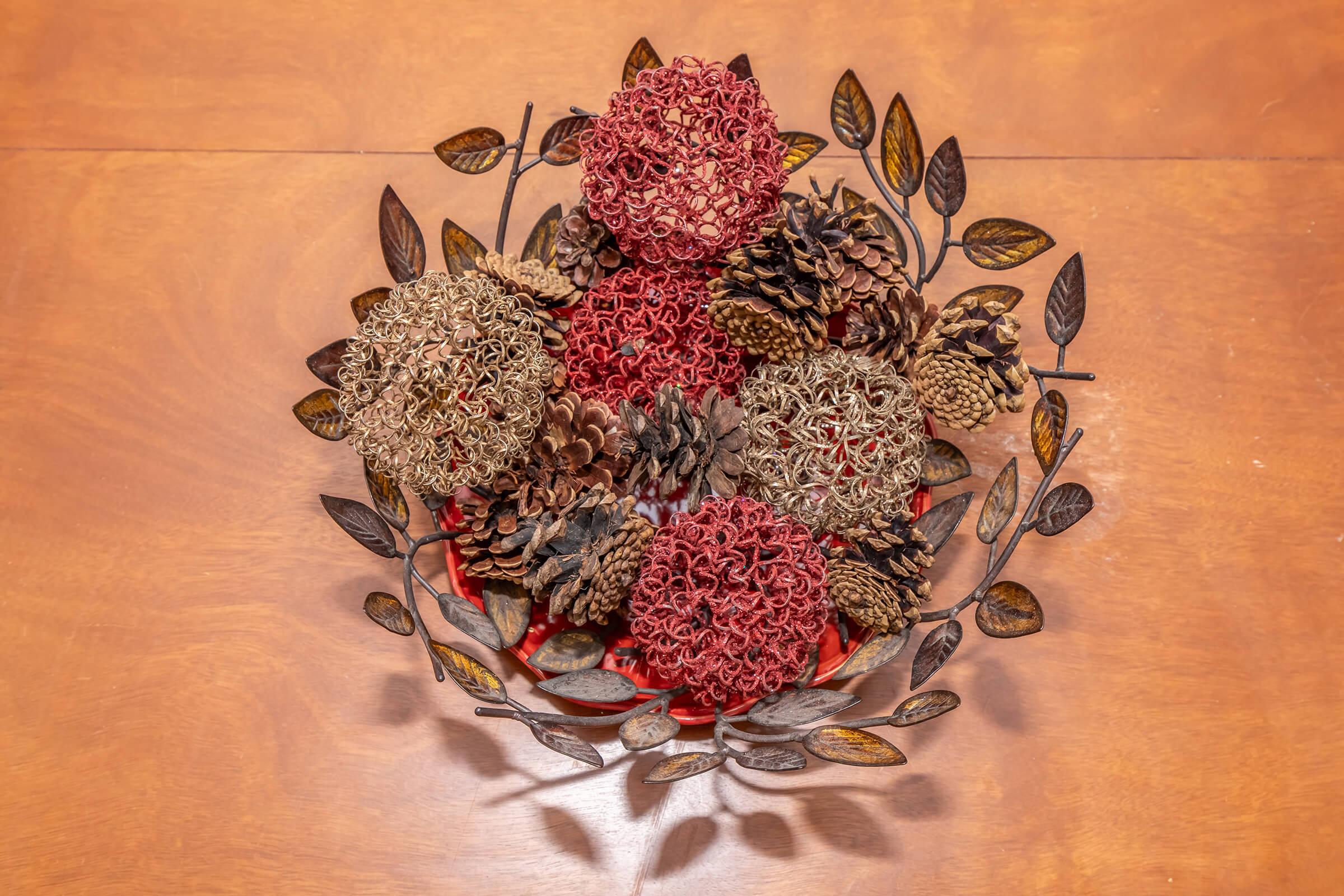 A decorative centerpiece featuring a variety of dried flowers and pinecones arranged in a circular metal bowl with leaves, set against a wooden surface. The colors include deep reds, browns, and natural tones, creating an inviting autumn aesthetic.