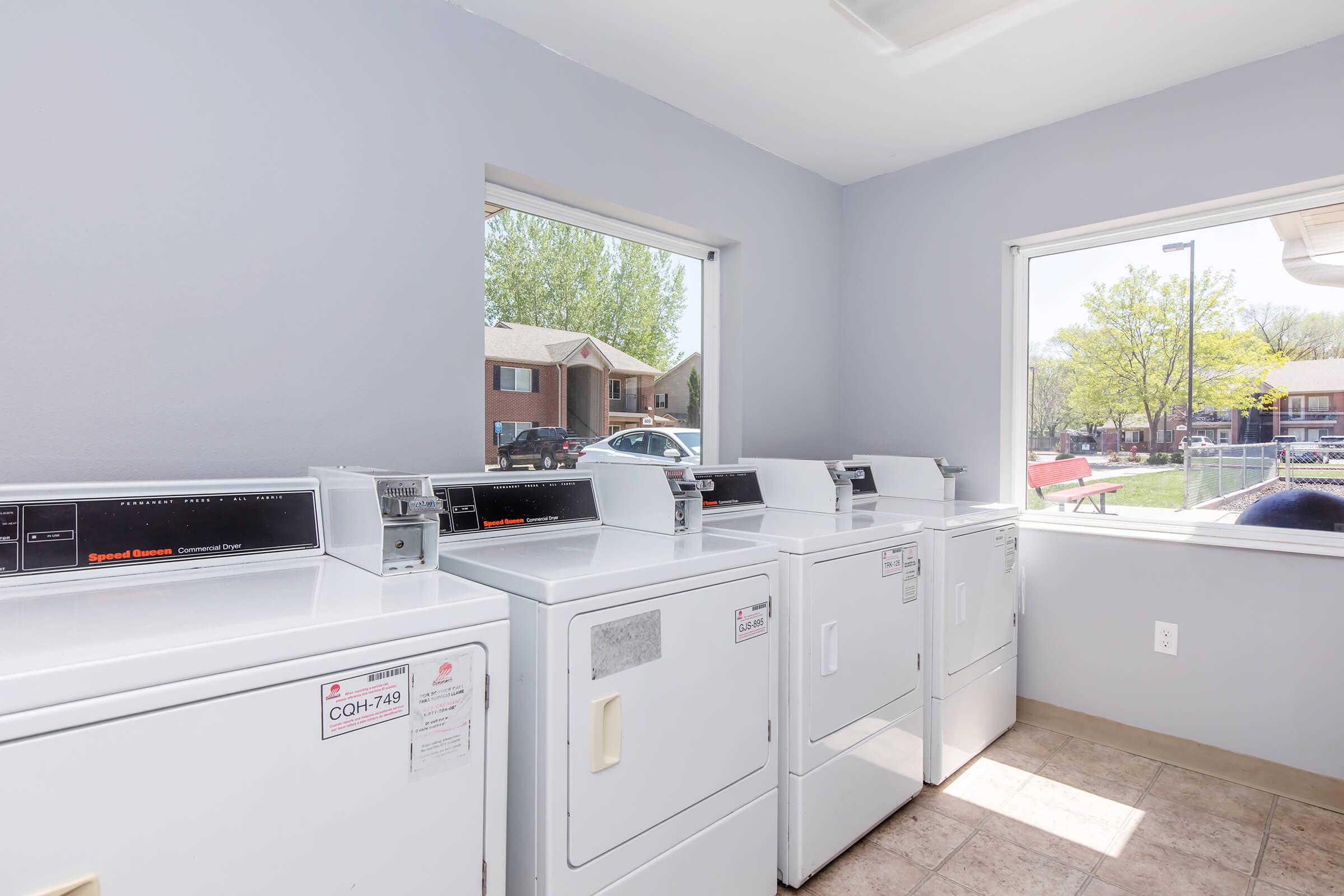 A laundry room featuring several white washing machines and dryers against a light gray wall. There are large windows providing natural light and a view of outdoor greenery and nearby buildings. The floor is tiled, and there are labels on the machines.