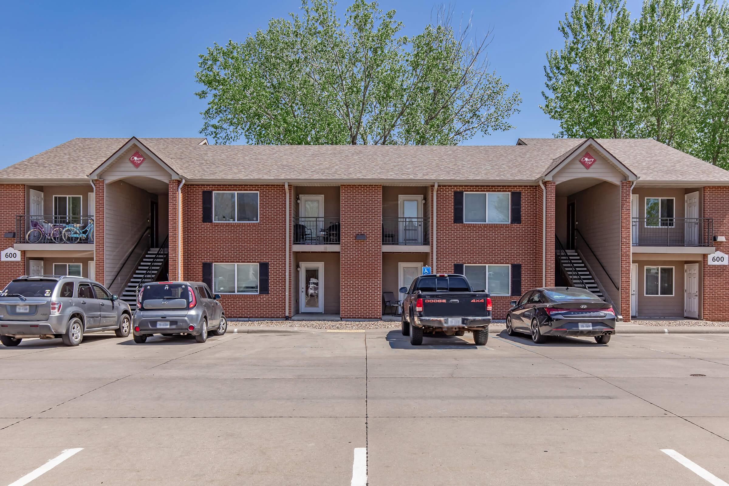 Two-story apartment building with a brick exterior and multiple units. The facade features large windows and balconies. Four parking spots are visible in front, occupied by two SUVs and a pickup truck. Green trees can be seen in the background under a clear blue sky.