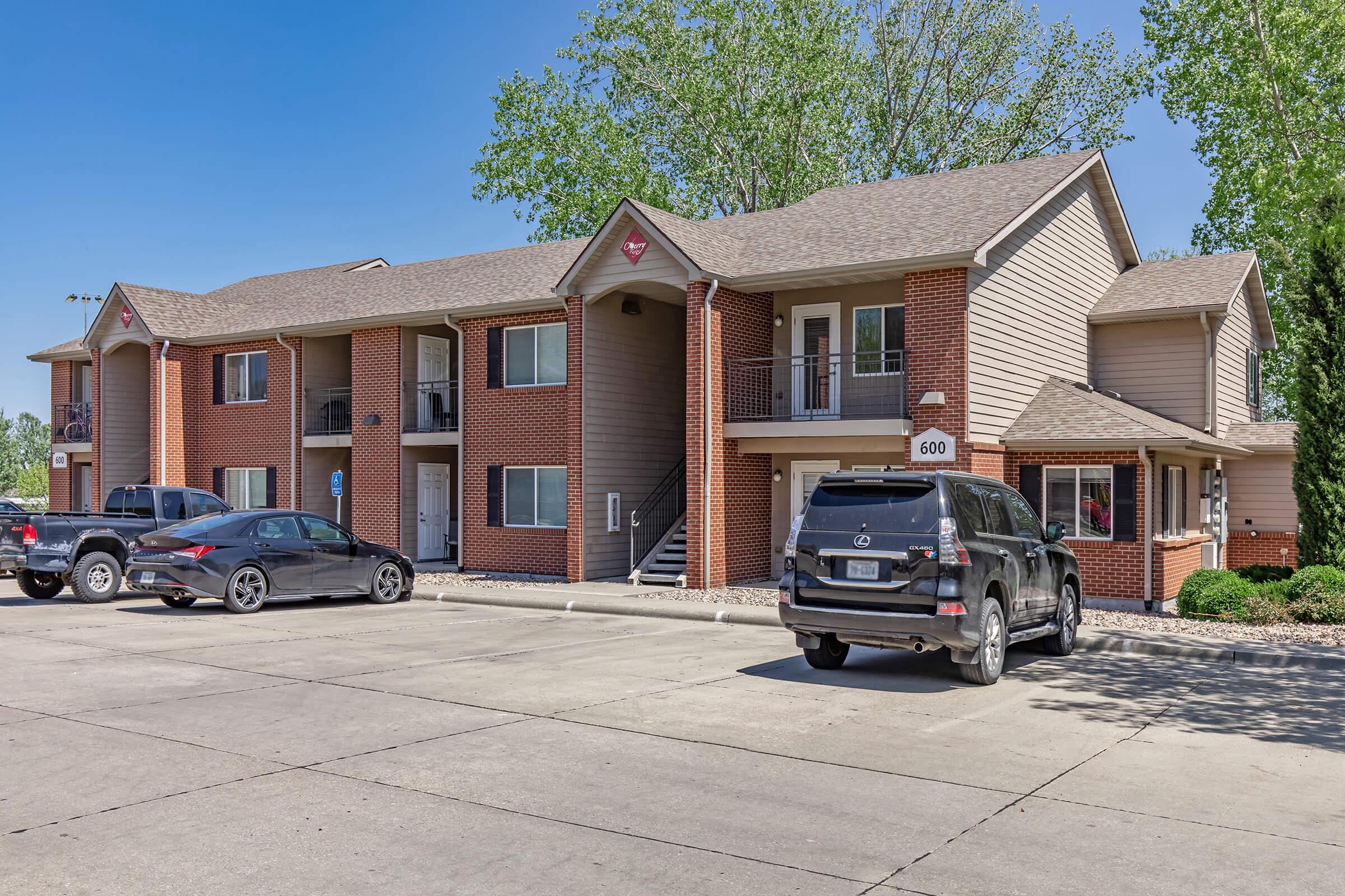 A two-story apartment building featuring red brick and tan siding, with multiple balconies. Several parked cars are visible in the foreground, and trees are seen in the background under a bright blue sky. The building has a well-maintained exterior and clear parking spaces.
