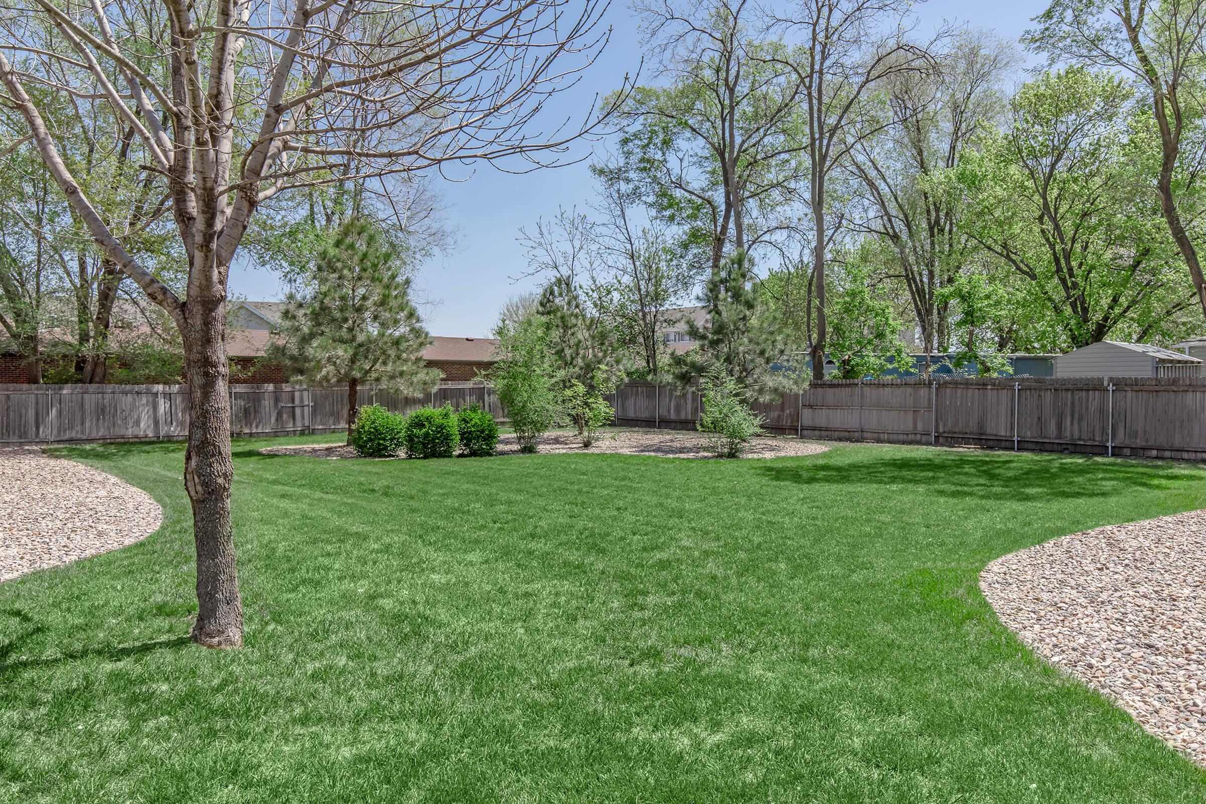 A lush green lawn surrounded by trees and shrubs in a backyard setting. The area features pathways made of gravel, with a wooden fence enclosing the space. The scene is bright and sunny, highlighting the greenery and landscaped features of the yard.