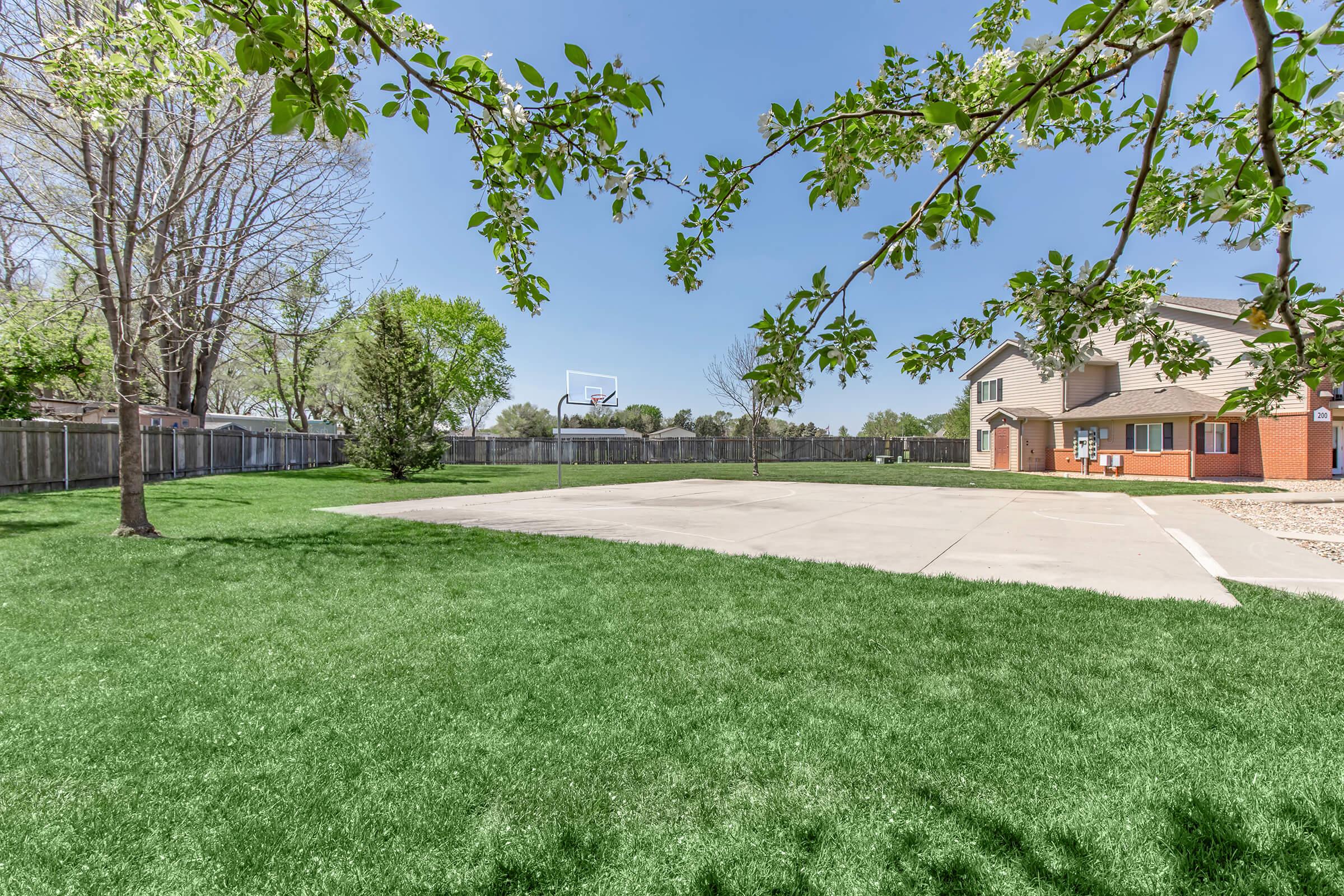 A spacious outdoor area featuring a paved basketball court surrounded by lush green grass and trees. In the background, there is a residential building with a fenced yard. The scene is set under a clear blue sky, suggesting a bright, sunny day.