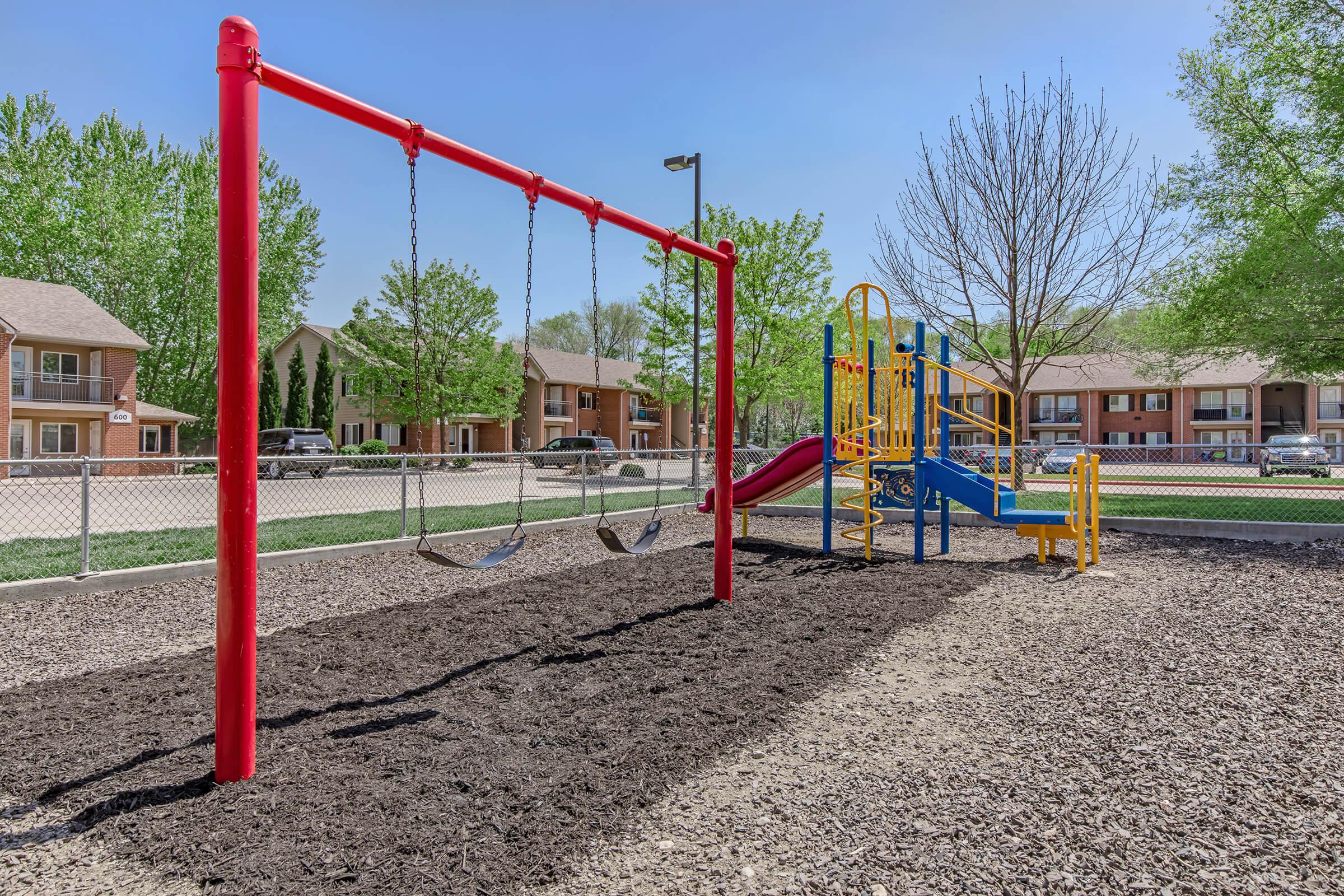 A colorful playground featuring a red swing set and a yellow and blue slide structure, surrounded by wood mulch. In the background, there are residential buildings and trees under a clear blue sky. The playground is fenced and well-maintained, providing a safe space for children to play.