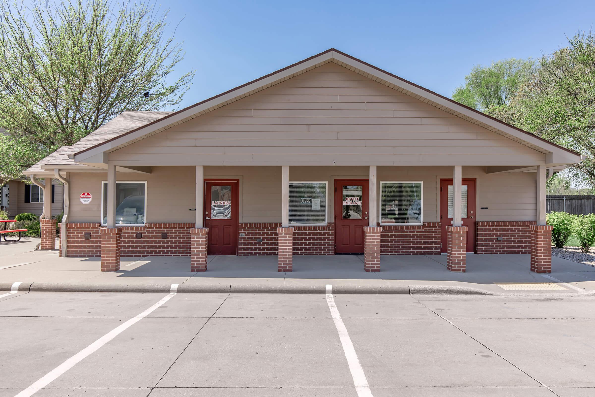 Single-story building with a beige exterior and red doors. It features multiple windows and is situated in a paved area with parking spaces. Surrounding greenery includes small trees and shrubs, creating a tidy appearance. The sky is clear and blue, indicating a sunny day.