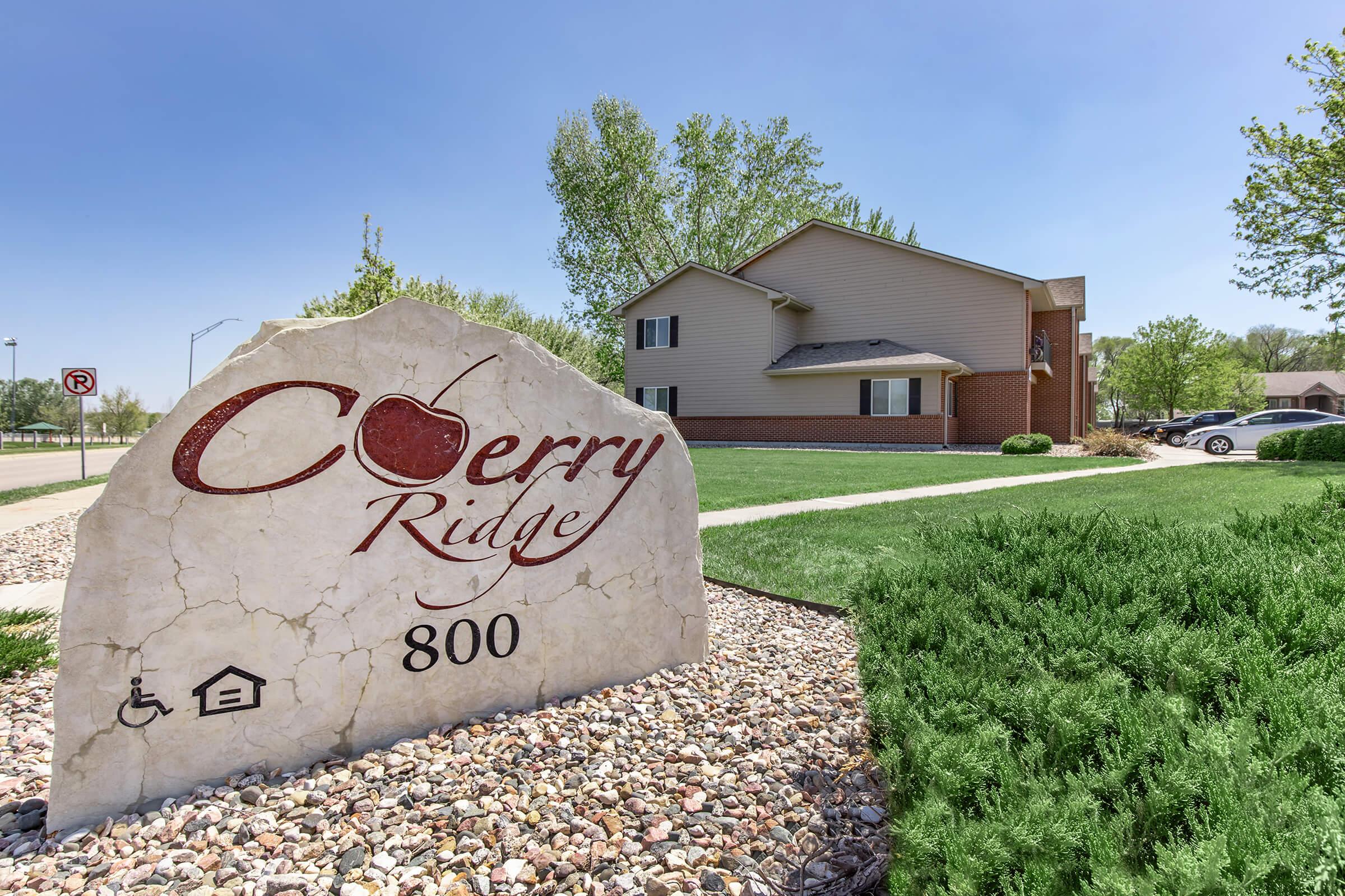 Sign for Cherry Ridge, displaying the number 800, surrounded by landscaped greenery and rocks. In the background, a residential building is visible, with clear blue skies above.