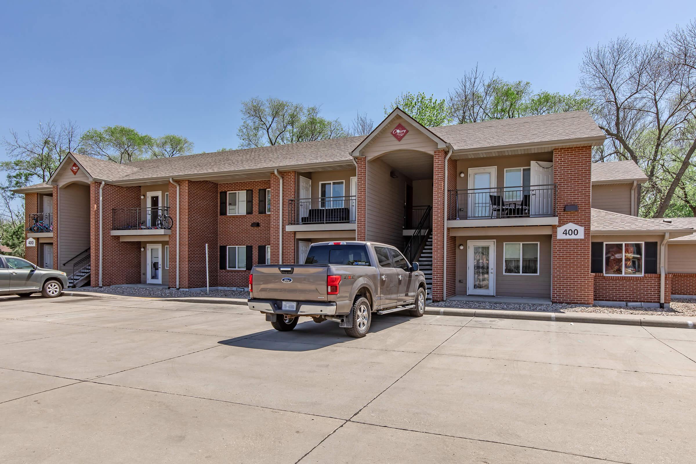 A two-story brick apartment building with balconies and a stone pathway. A gray pickup truck is parked in front, and trees can be seen in the background. The building features a sign indicating the unit number "400." The area appears well-maintained and landscaped.