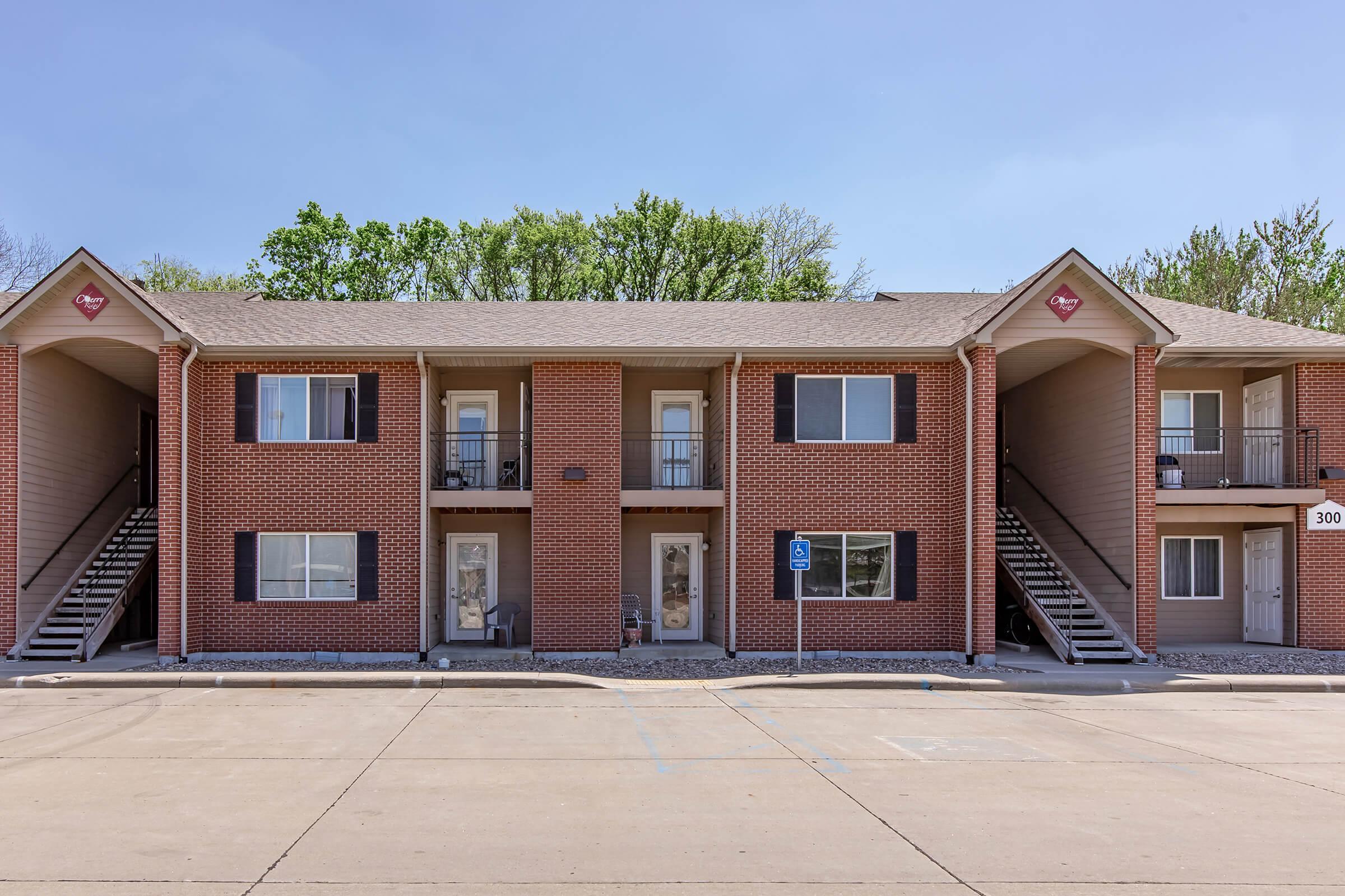 Two-story brick apartment building with multiple units, featuring stairs on either side. The facade includes dark shutters and number plaques by the doors. A parking lot is visible in front with a designated handicap space. Green trees can be seen in the background under a clear blue sky.