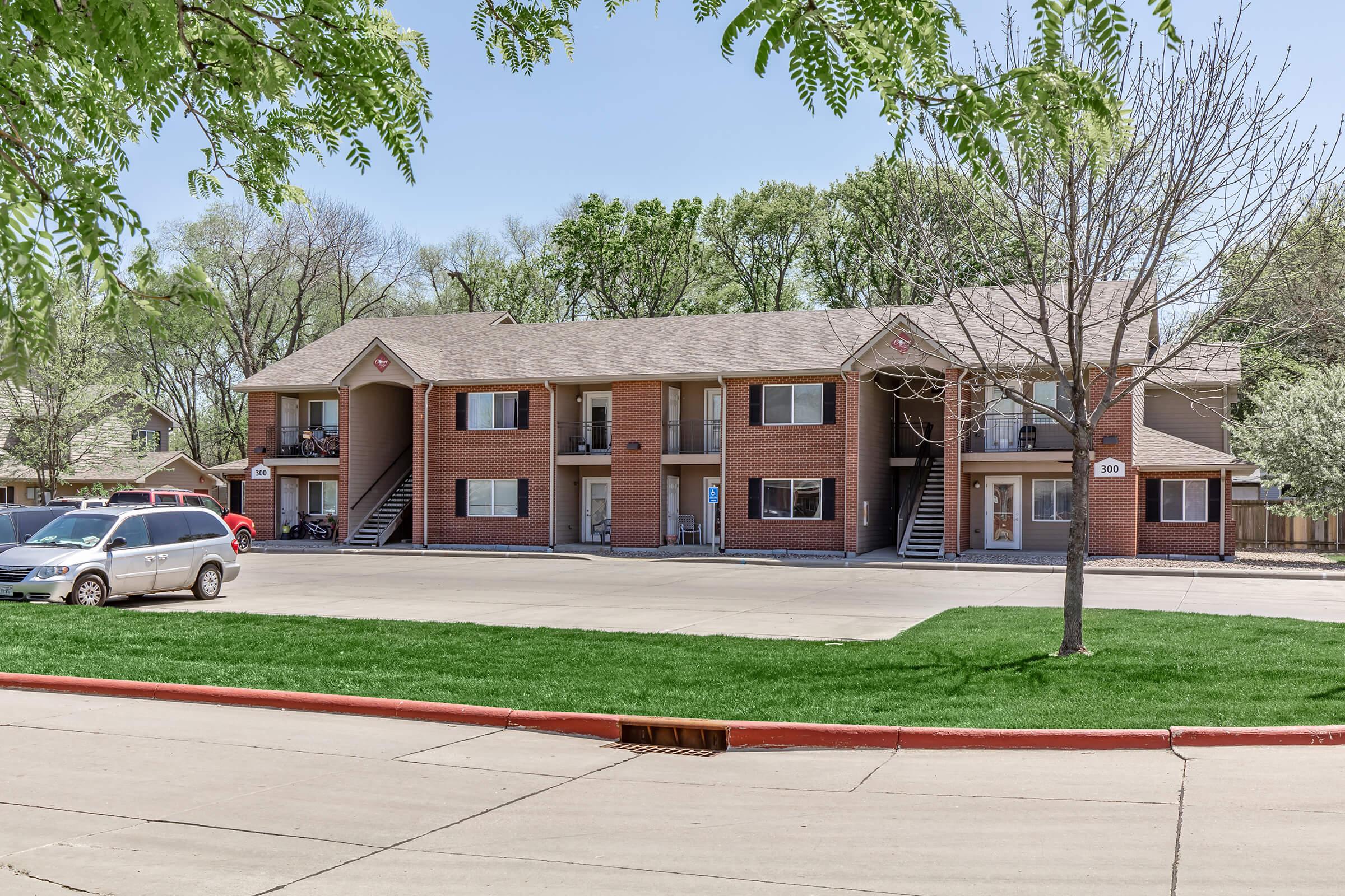 A well-maintained two-story apartment building surrounded by green grass and trees. The building features multiple entrances with stairs leading to each unit. A parked car is visible in the foreground, and the area is well-lit with clear blue skies.