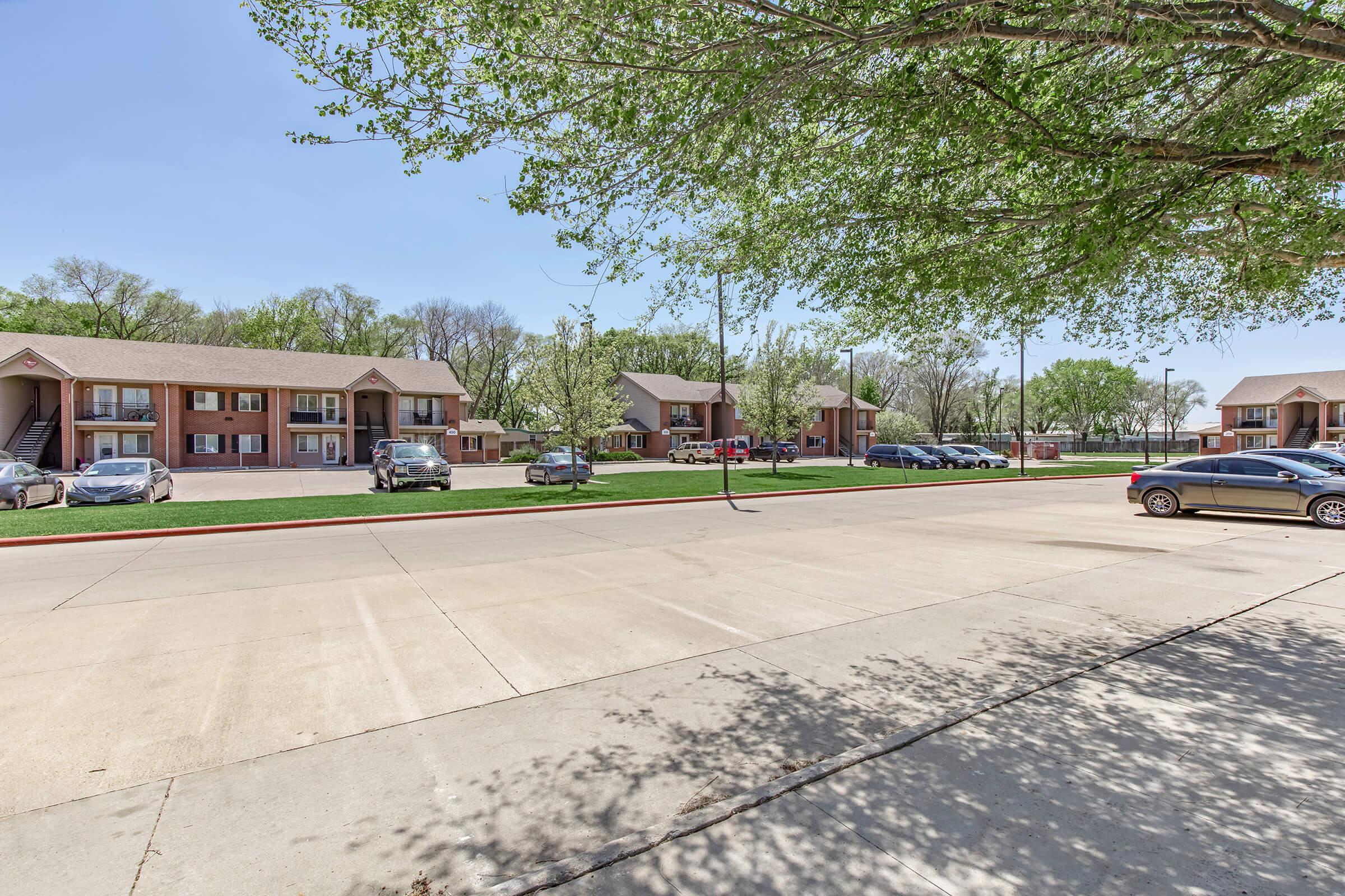 A view of a residential area featuring two-story apartment buildings surrounded by green trees. A paved driveway runs in the foreground with parked cars on either side. The scene is bright and sunny, showcasing a well-maintained landscape.