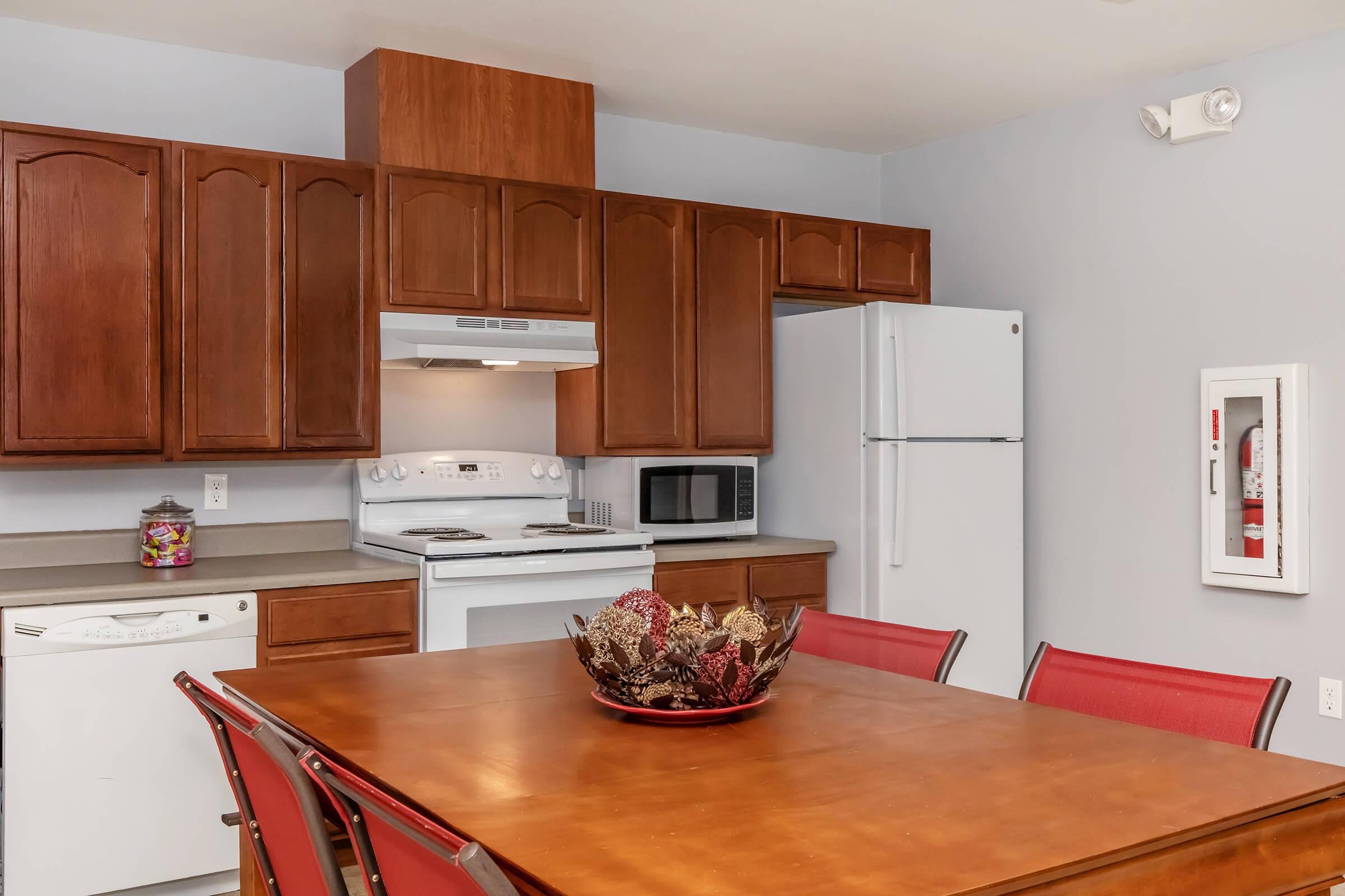 A well-equipped kitchen featuring wooden cabinets, a white refrigerator, microwave, and stove. The table in the center is set with a decorative bowl of flowers and a multi-colored centerpiece. The walls are painted a light gray, enhancing the modern, functional space.