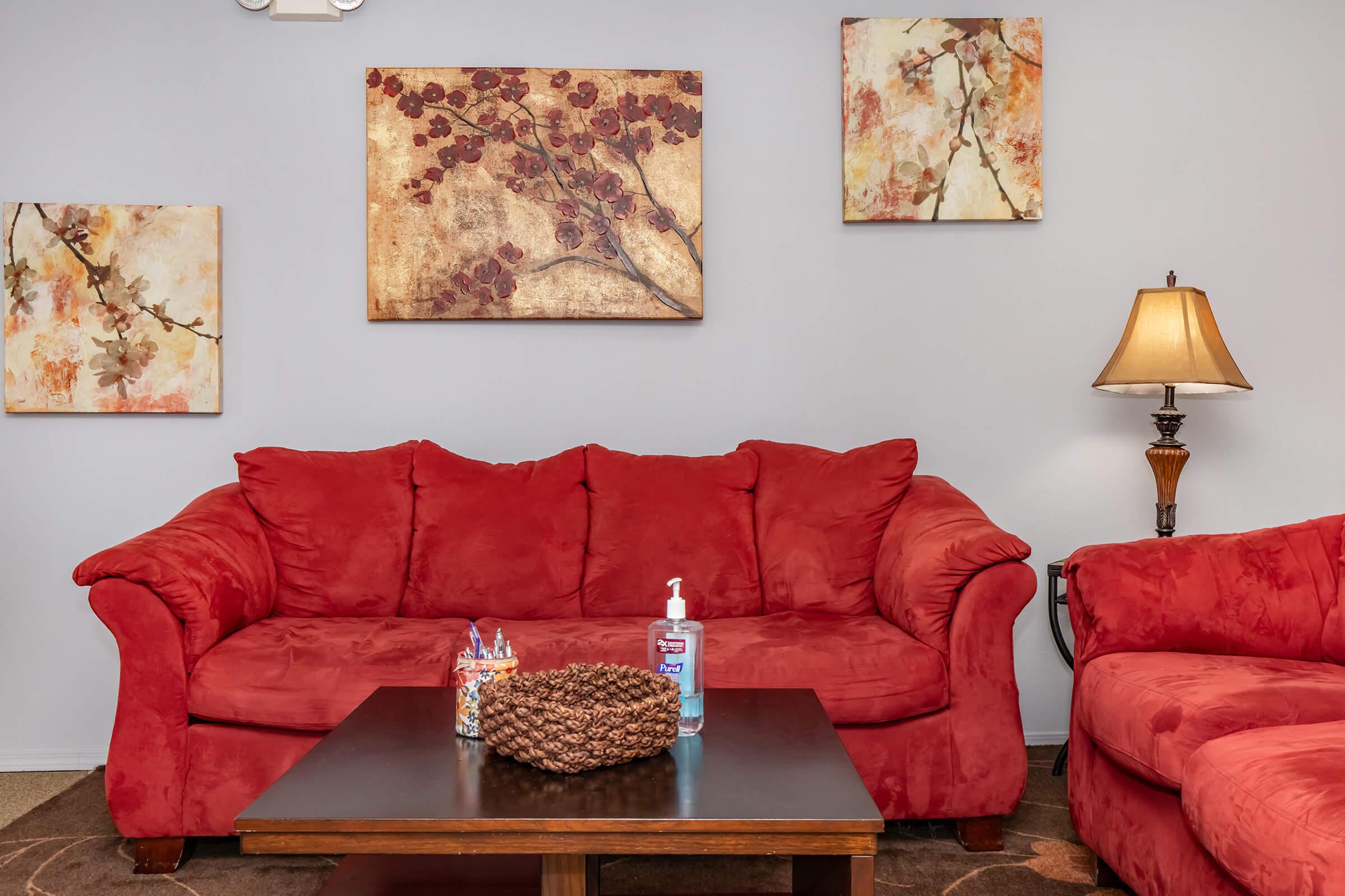 A cozy living room featuring a red couch and armchair, a wooden coffee table with a decorative pinecone and hand sanitizer, and two abstract floral wall art pieces. A lamp adds warm lighting to the space.