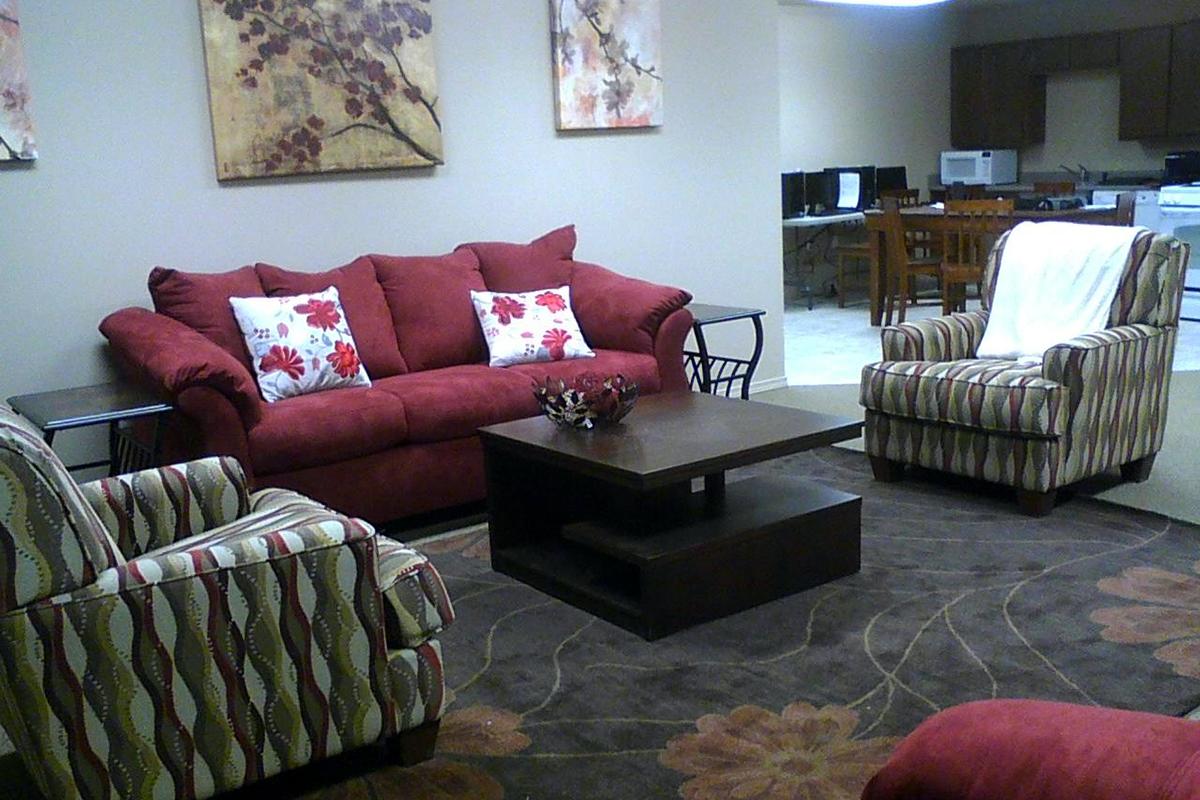 A cozy living area featuring a red sofa with floral pillows, two striped armchairs, and a coffee table. The floor is covered with a patterned rug, and there are decorative wall art pieces above the couch. In the background, a glimpse of a kitchen area with appliances and a dining table can be seen.