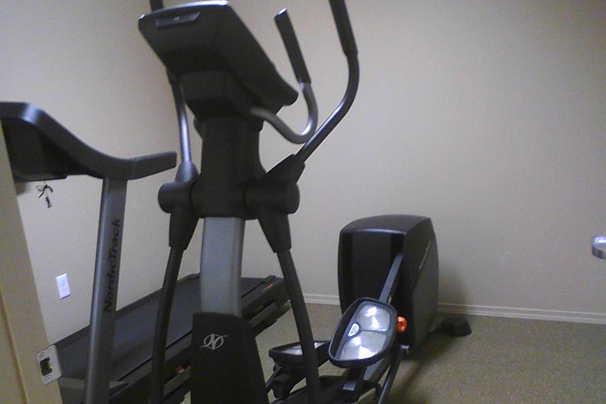 A home gym setting featuring an elliptical trainer and a treadmill against a neutral-colored wall. The equipment is positioned closely together, emphasizing a compact workout space. Soft lighting highlights the modern design of the machines.