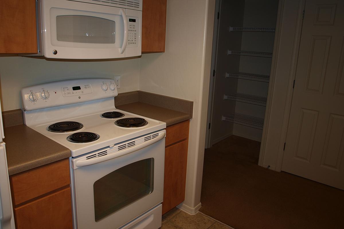 A view of a kitchen featuring a white electric stove with four burners, a microwave above, and wooden cabinets. To the right, there's an open door leading to a pantry with shelves, while the floor is a light-colored tile and the kitchen has beige walls.