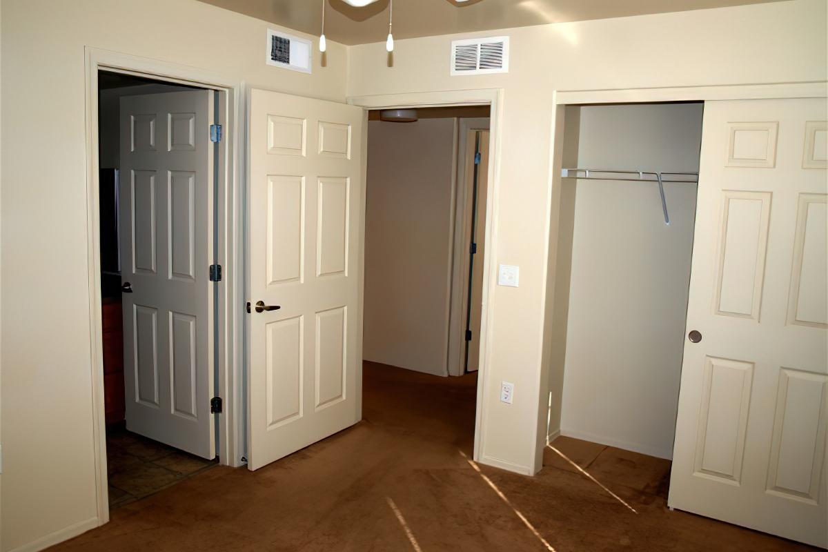 A well-lit interior space featuring two closed doors and an open closet. The walls are painted beige, and the floor is carpeted in a light brown shade. The ceiling has a ceiling fan and overhead lighting, creating a cozy and inviting atmosphere.