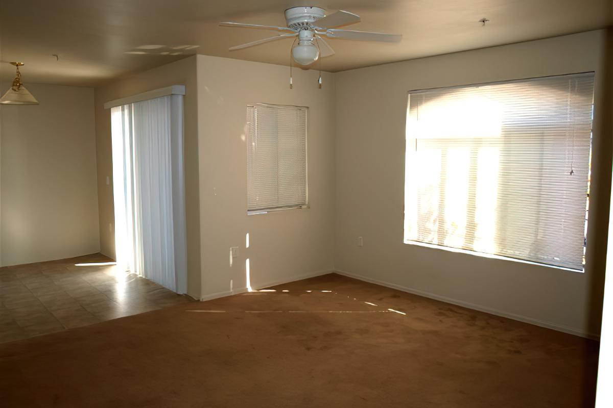 A spacious, empty living room featuring beige walls and carpeting. There are windows with blinds allowing natural light to filter in. A ceiling fan is installed, and a light fixture hangs from the ceiling. The room appears clean and ready for furnishings.