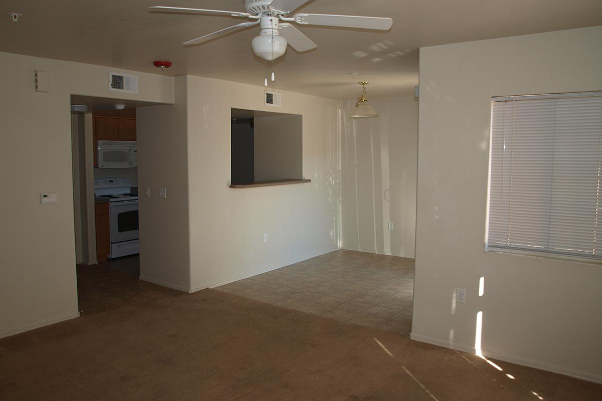 A spacious, empty living room with light brown carpet, a ceiling fan, and a window with blinds. An adjacent kitchen is partially visible with white appliances. The walls are painted a light color, and there is a pendant light hanging from the ceiling. The overall atmosphere is bright and uncluttered.