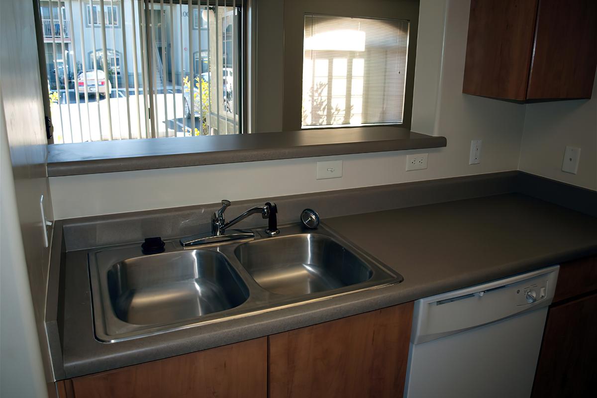 A modern kitchen sink area featuring a double-basin stainless steel sink, a chrome faucet, and a dishwasher below the counter. The countertops are gray and the cabinetry is a warm wood finish. A window above the sink provides natural light, with a view of the outside.