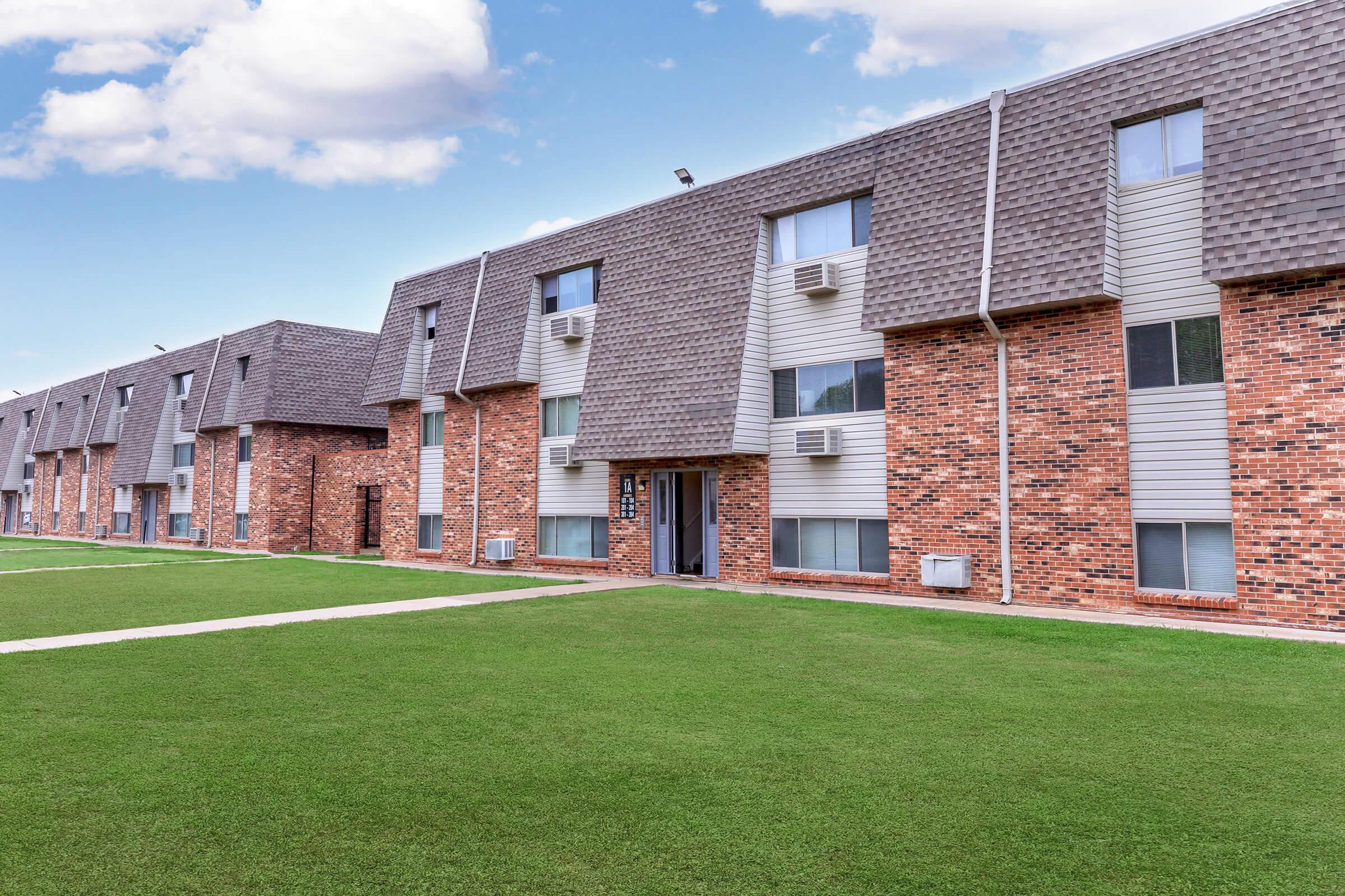 A row of brick apartment buildings with pitched roofs, featuring multiple windows and air conditioning units. A well-maintained green lawn runs in front, with a clear blue sky and scattered clouds above, creating a pleasant residential atmosphere.